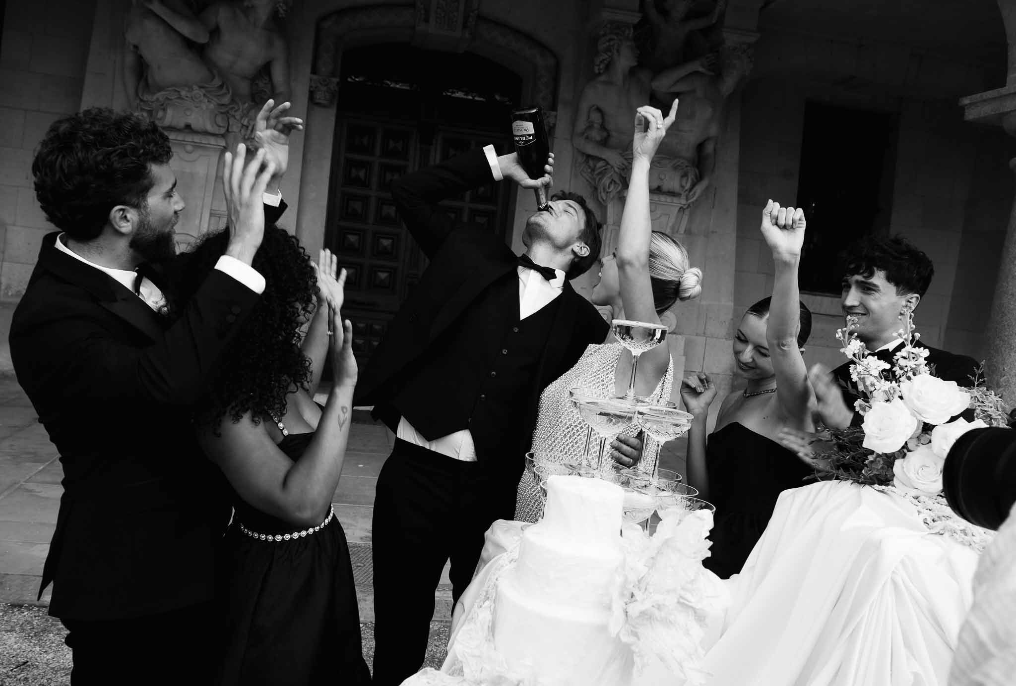 Black and white candid of groom drinking from champagne bottle at coupe tower with cheering guests