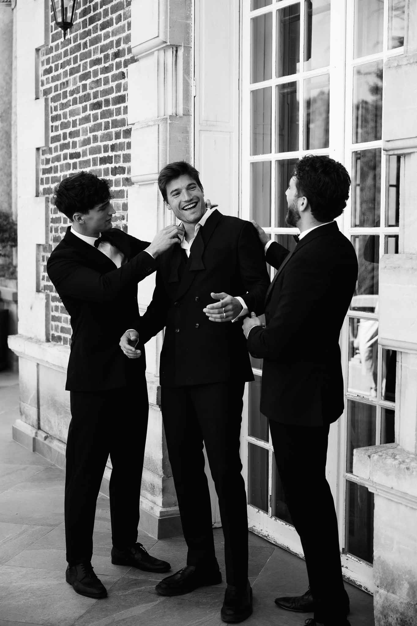 Black-and-white photo of groom laughing as two groomsmen adjust his suit outside a French building