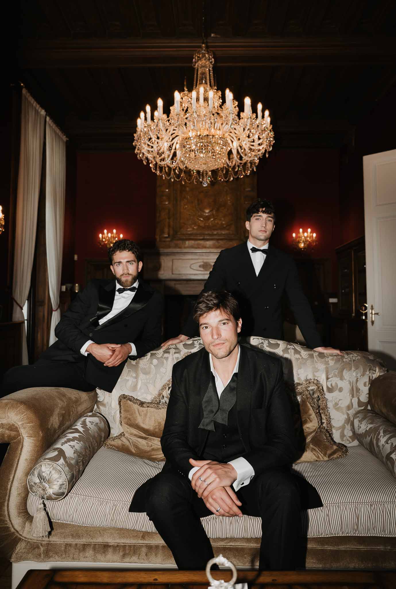 Groom and two groomsmen in black tuxedos on gold damask sofa in crimson chateau salon with crystal chandelier