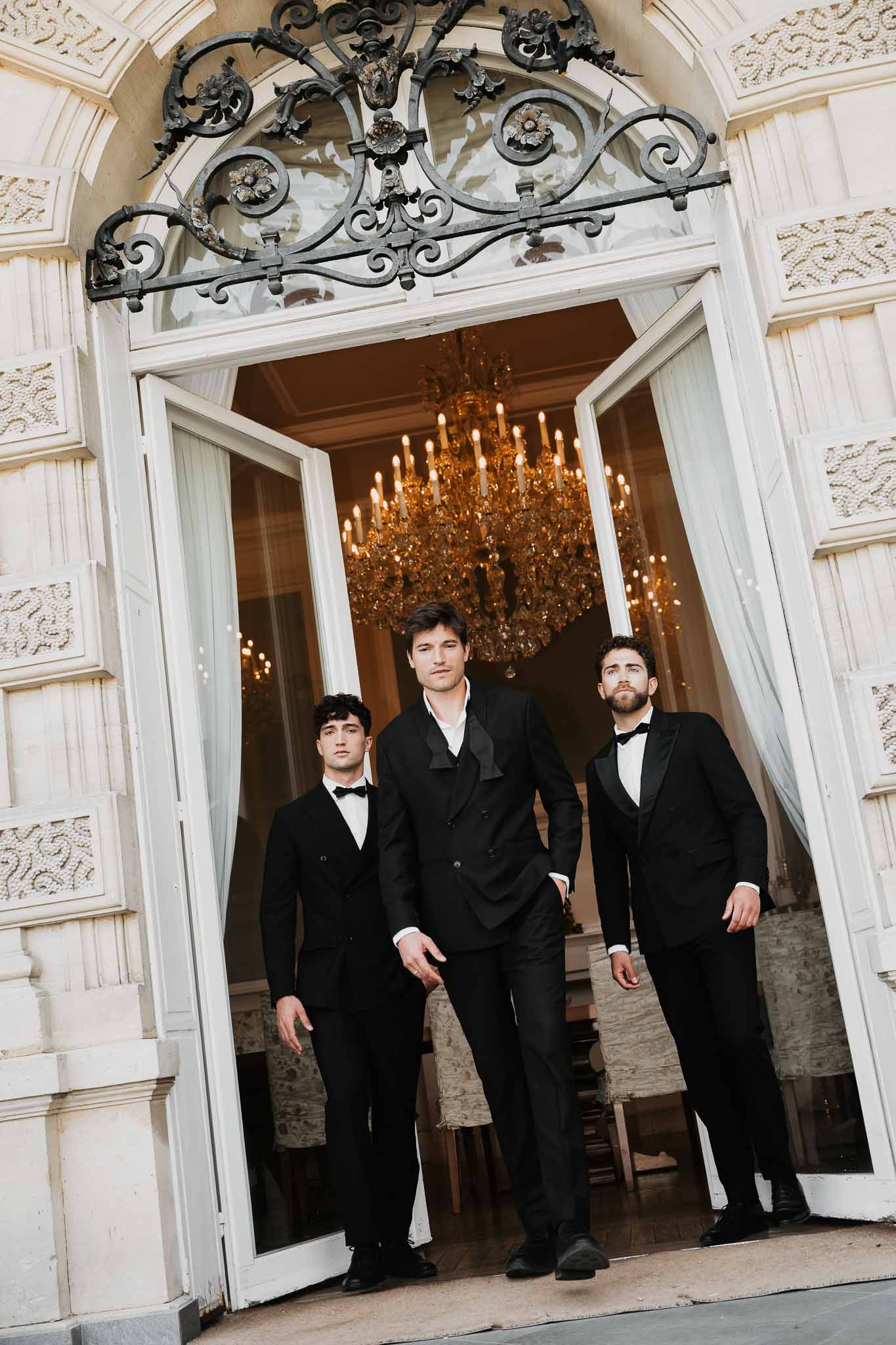Groom and two groomsmen in black suits stepping through ornate French doors with chandelier behind