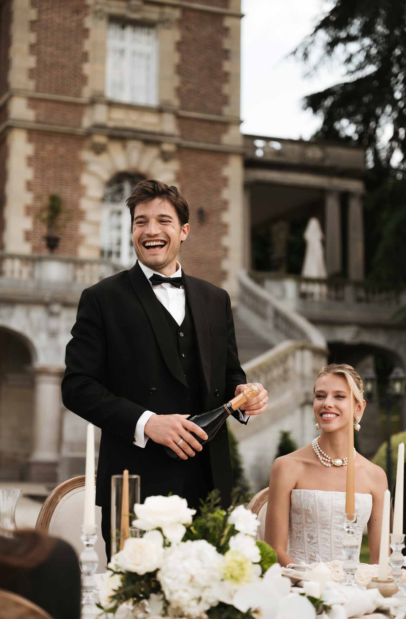 Groom holding champagne bottle mid-toast beside seated bride at peony-dressed table before brick chateau