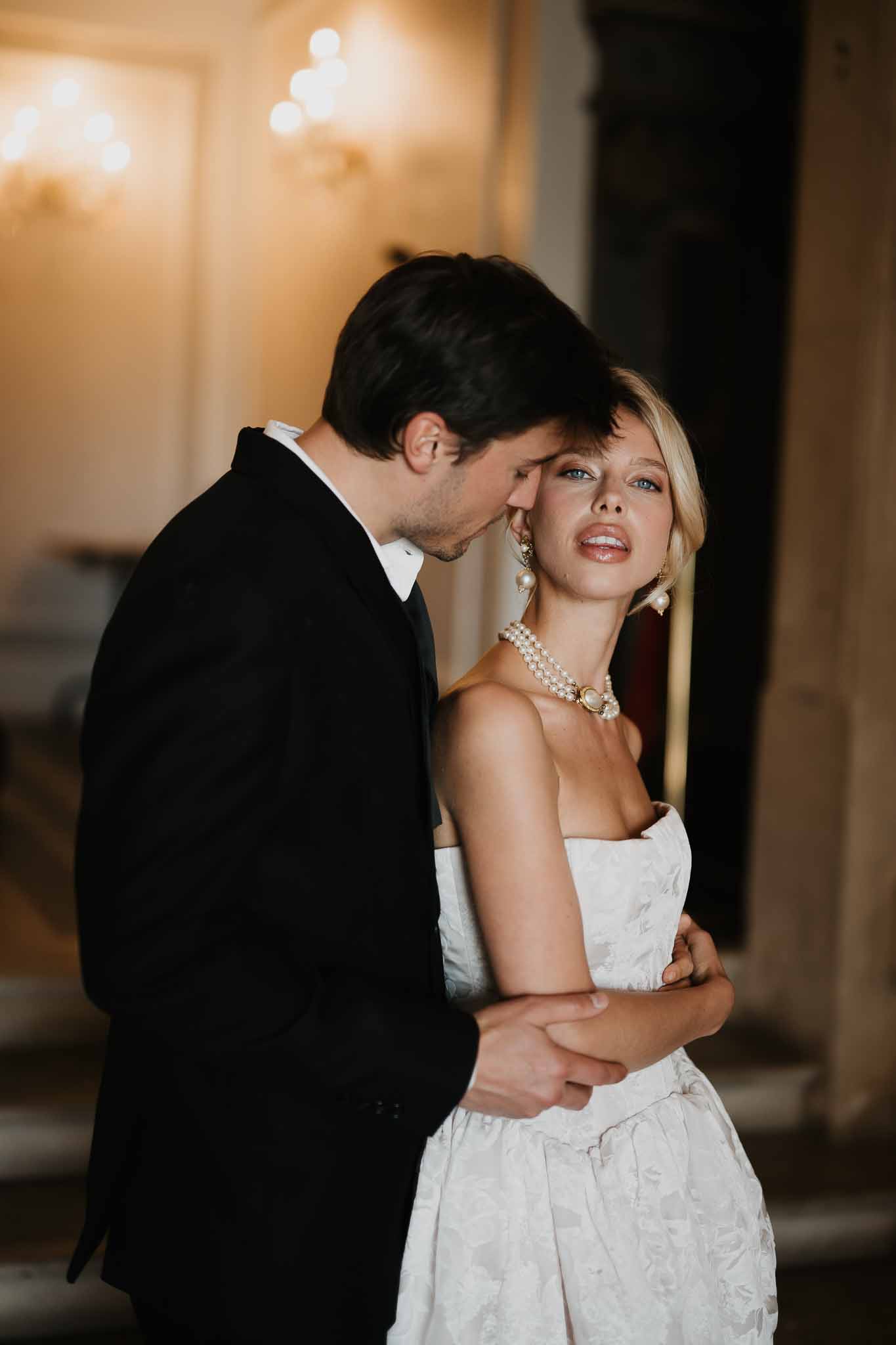 Groom kissing bride cheek in chandelier-lit chateau hall with pearl choker and jacquard gown