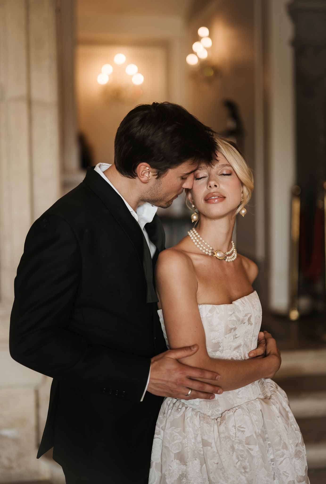 Groom in black tuxedo kissing bride's temple as she wears strapless floral jacquard gown and pearl choker necklace