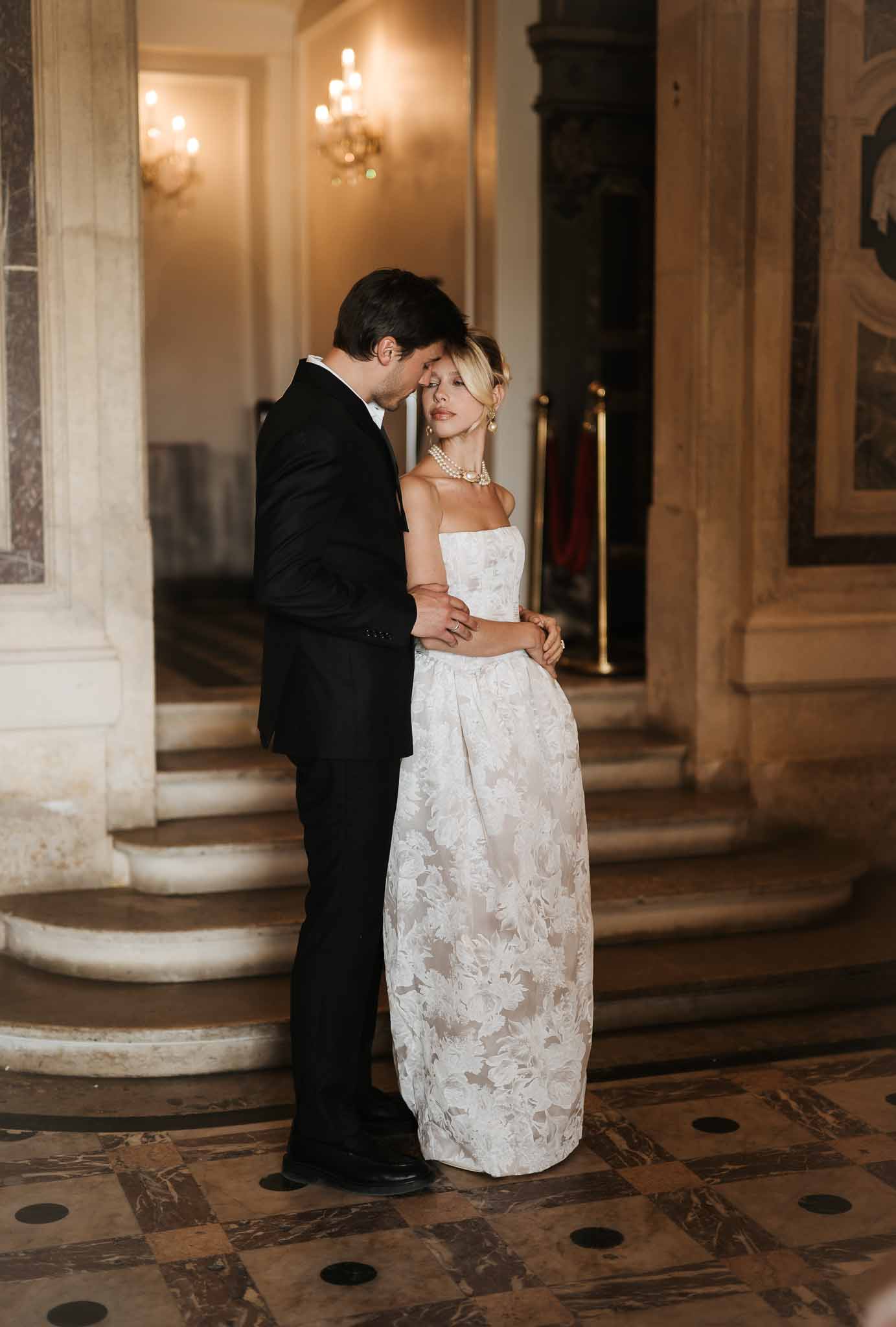 Groom embracing bride in strapless ivory jacquard gown with pearl necklace inside ornate marble-floored palace interior