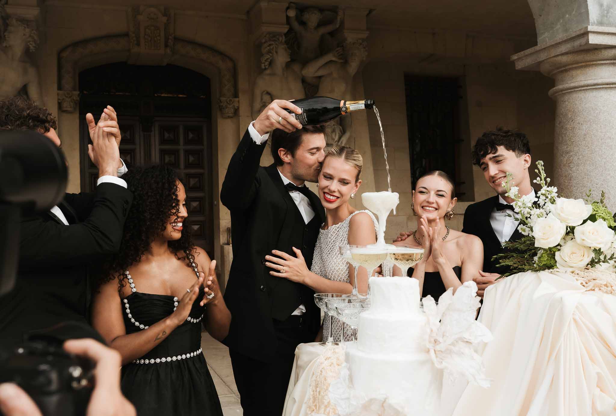 Groom pouring champagne tower and kissing bride beside white cake at chateau with clapping guests