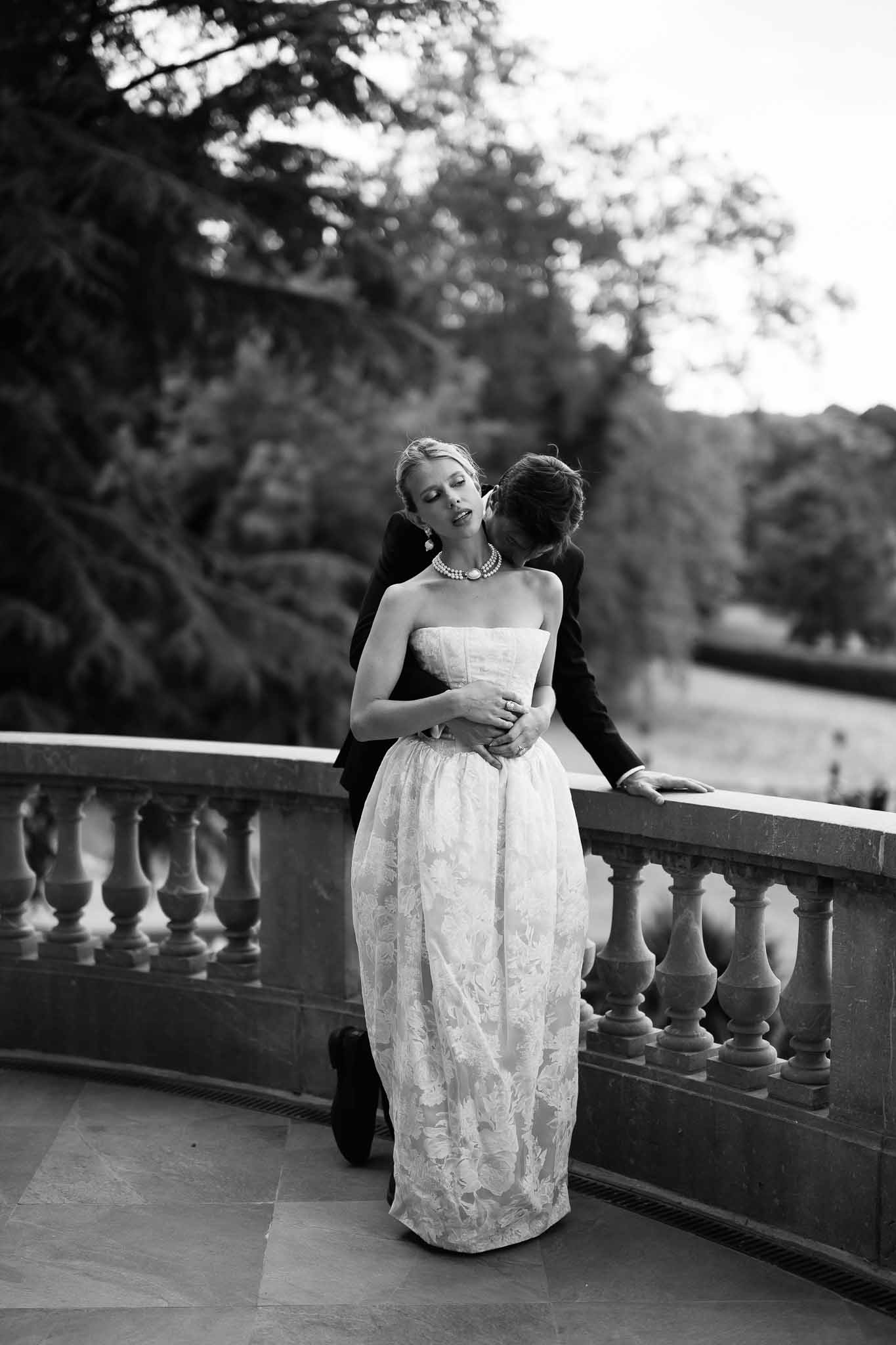 Black and white portrait of groom embracing bride on stone balustrade terrace with parkland behind