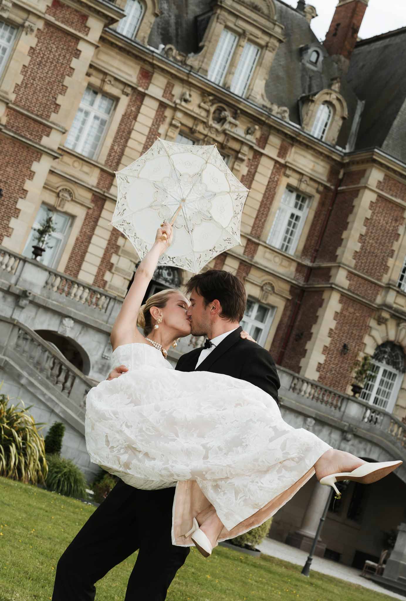 Groom carrying bride with lace parasol in front of brick chateau with mansard roof and balustrade