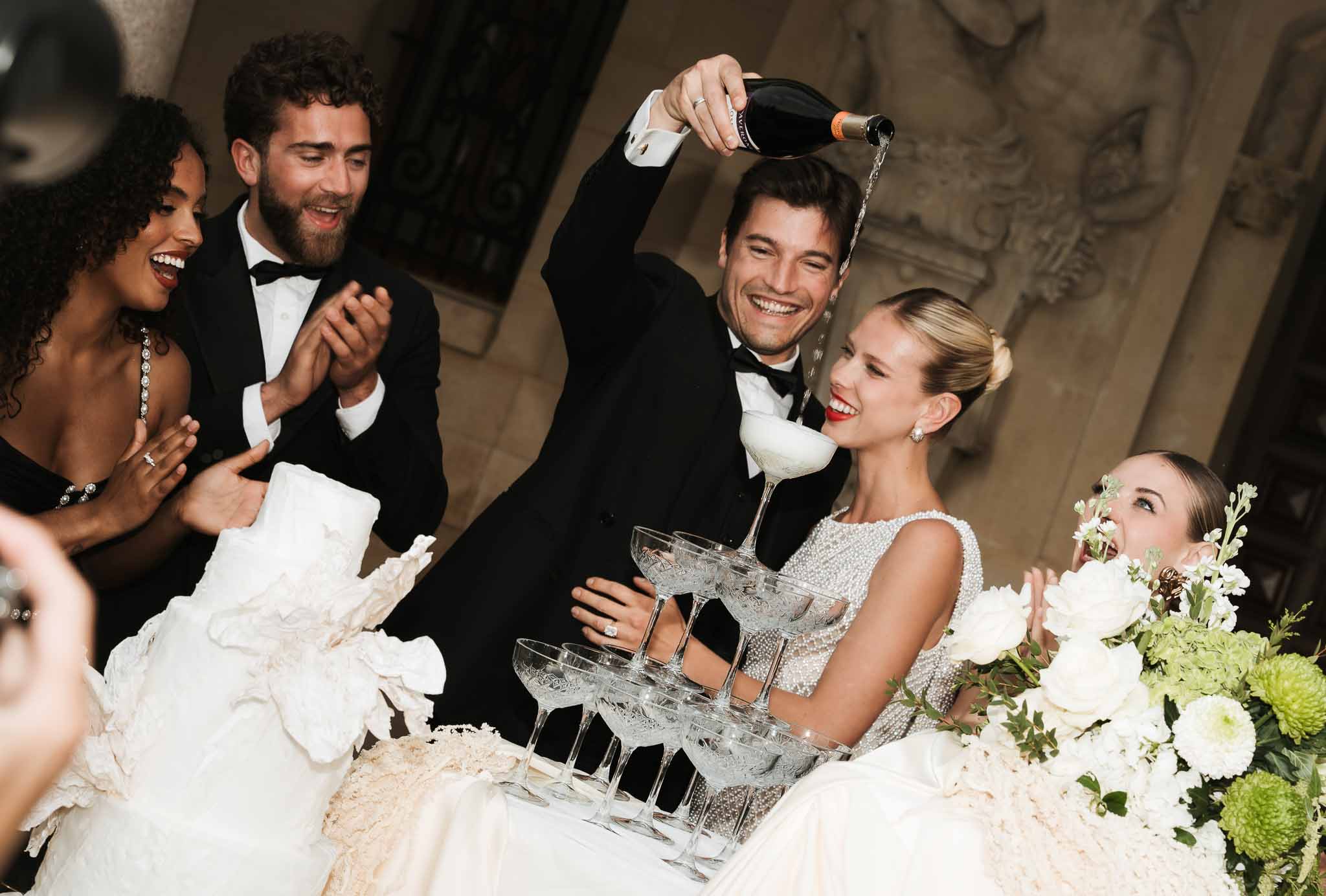 Groom pouring champagne into coupe tower as bride in pearl gown laughs with guests applauding beside tiered cake