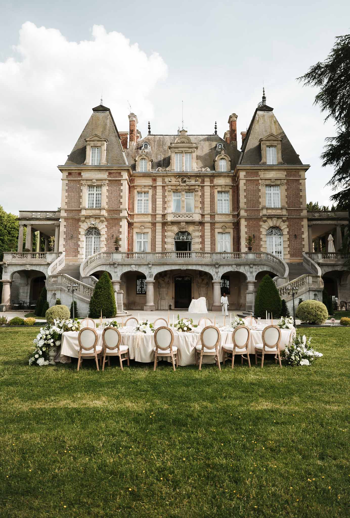 Long dining table with white hydrangea centerpieces and gold candle holders on lawn before grand French chateau