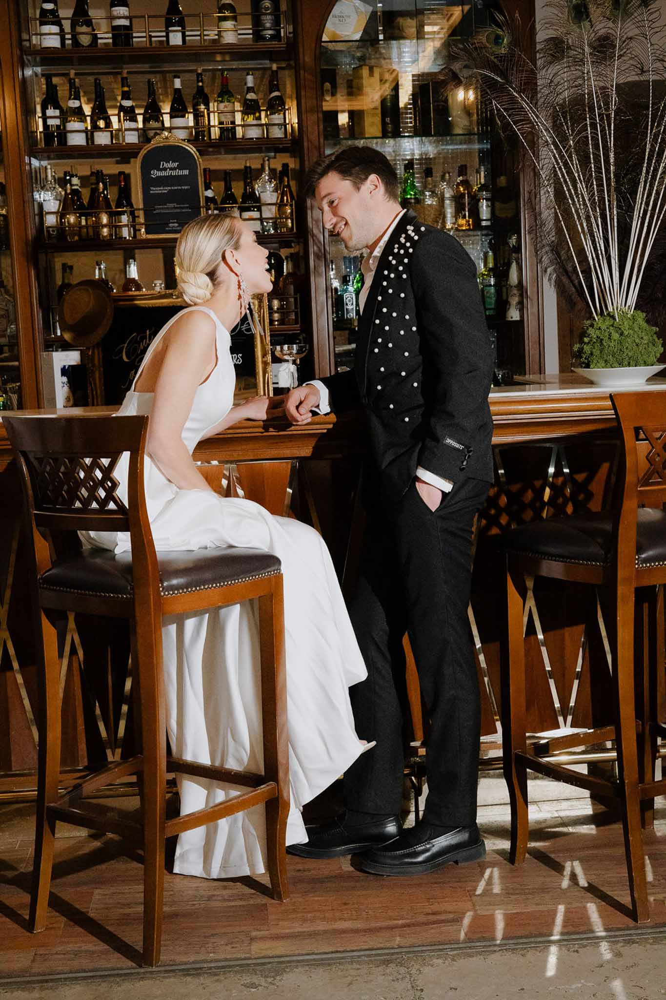 Bride and groom laughing at dark wood bar with wine bottles and dried pampas during reception