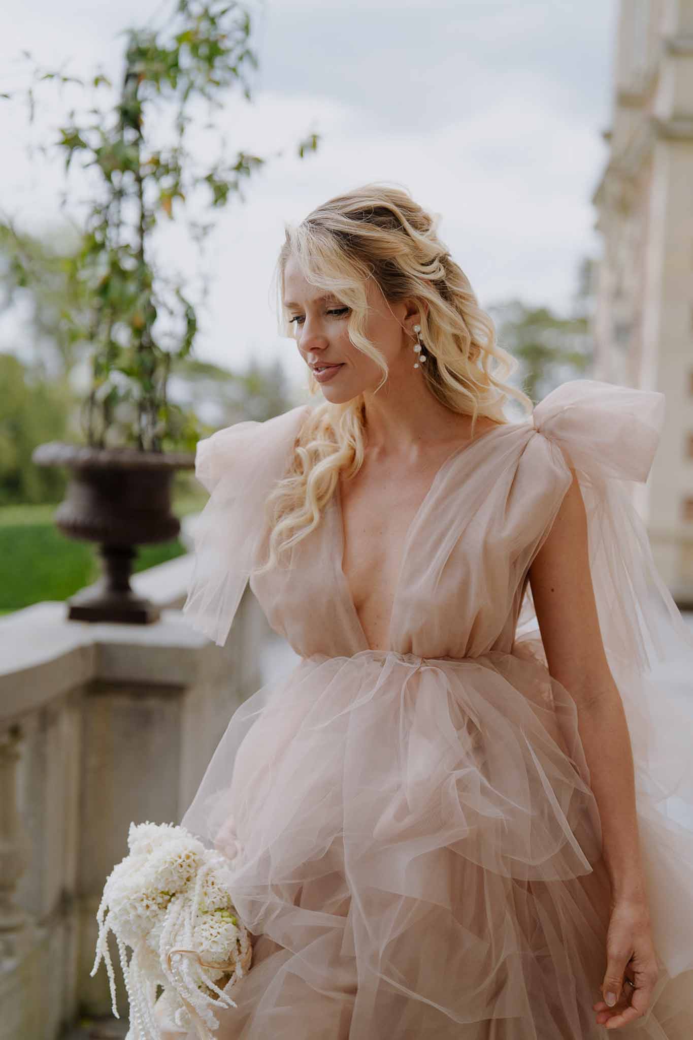 Bride in blush-pink ruffled tulle gown with bow shoulders holds ivory bouquet on chateau stone terrace