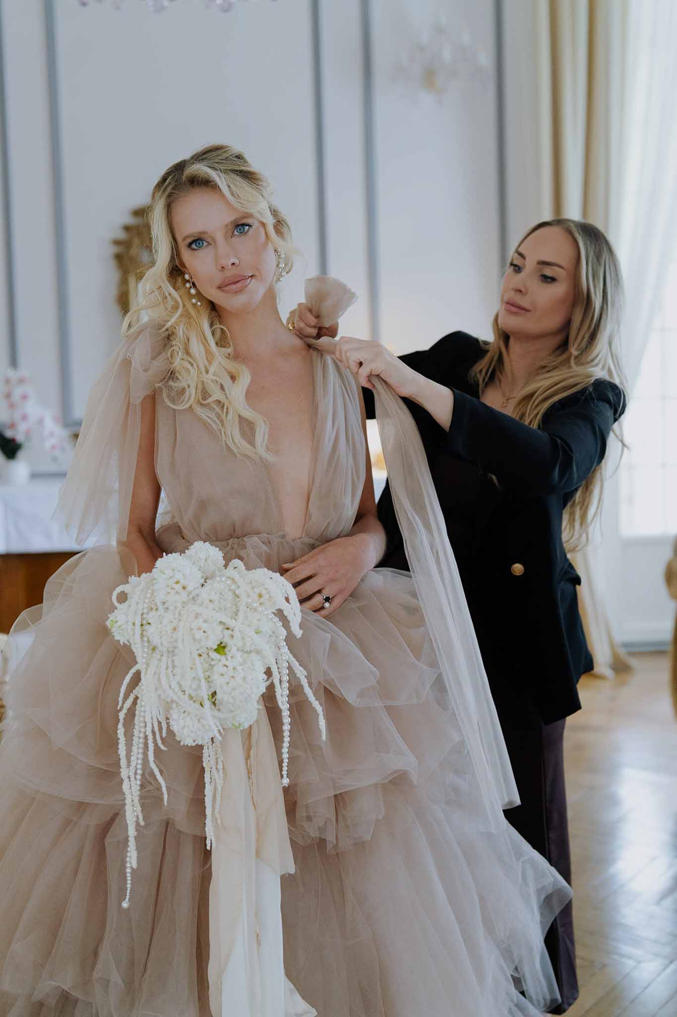 Bride in blush tulle ballgown holding white hydrangea bouquet while stylist adjusts her veil in paneled room