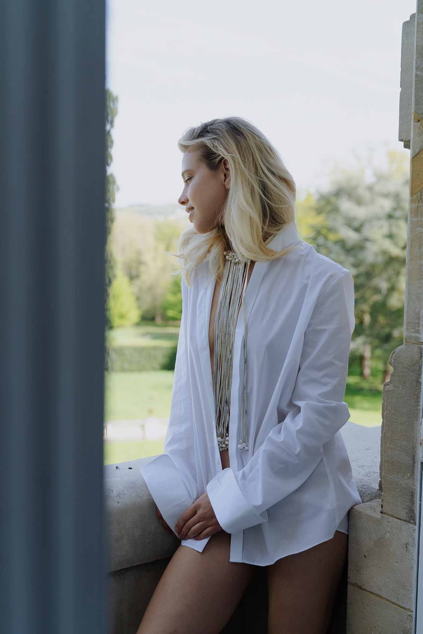 Bride in white shirt seated on chateau stone window ledge wearing layered silver and pearl statement necklace