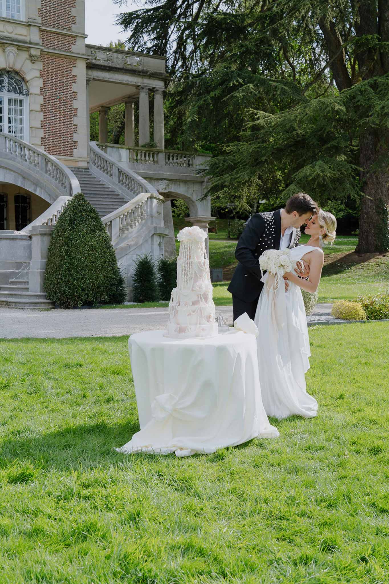 Couple kissing beside pearl-draped blush cake before chateau double staircase with topiary