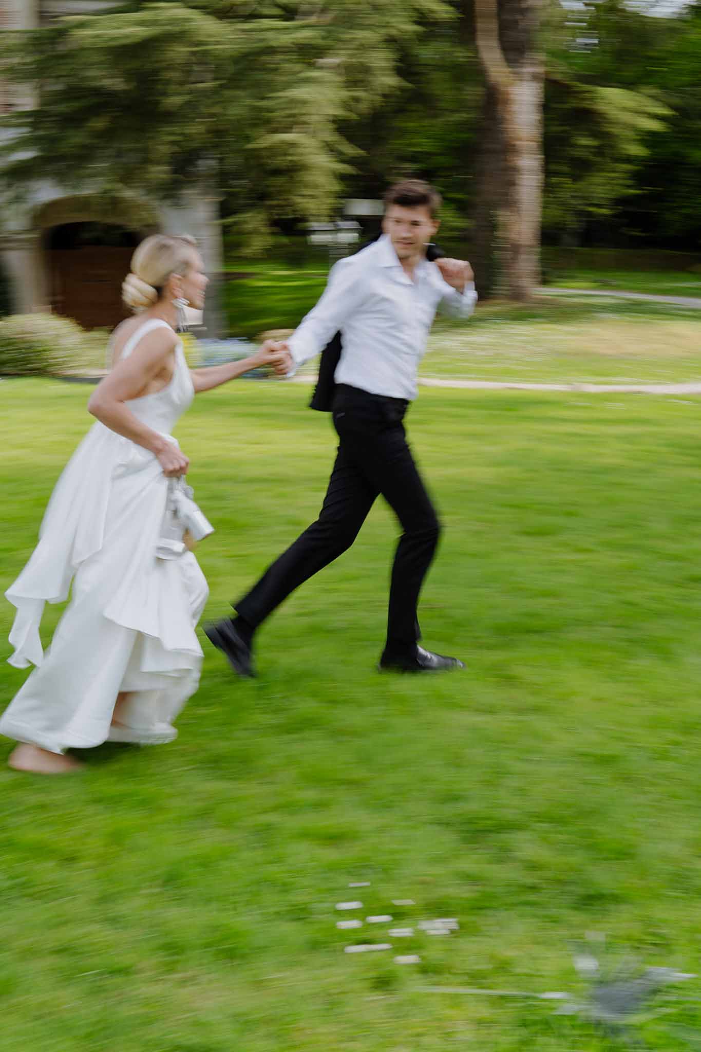 Motion-blurred couple running hand in hand across estate lawn with tiered skirt and removed jacket