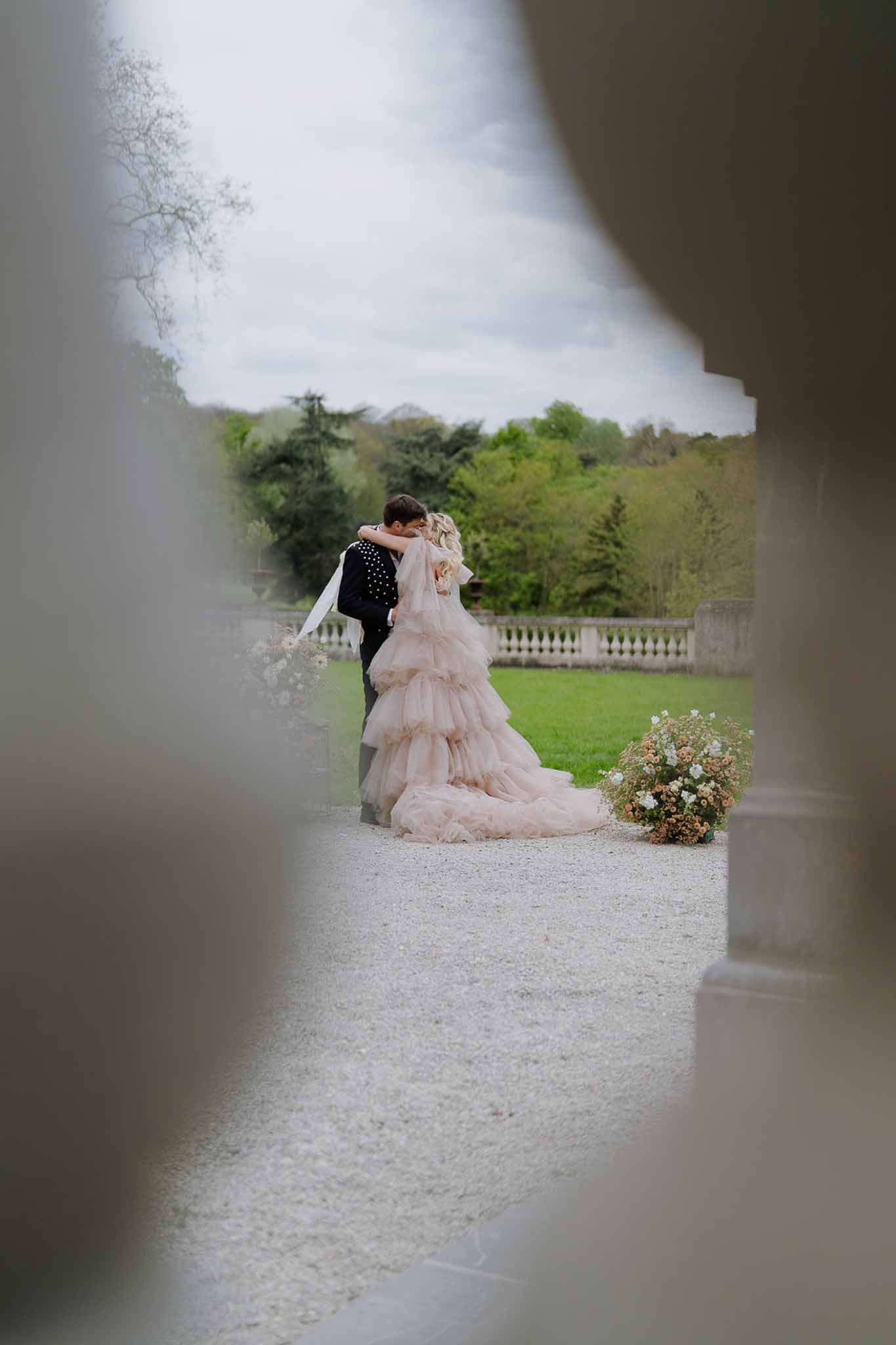 Bride in blush pink ruffle ballgown embracing groom in embellished jacket framed through stone columns at chateau