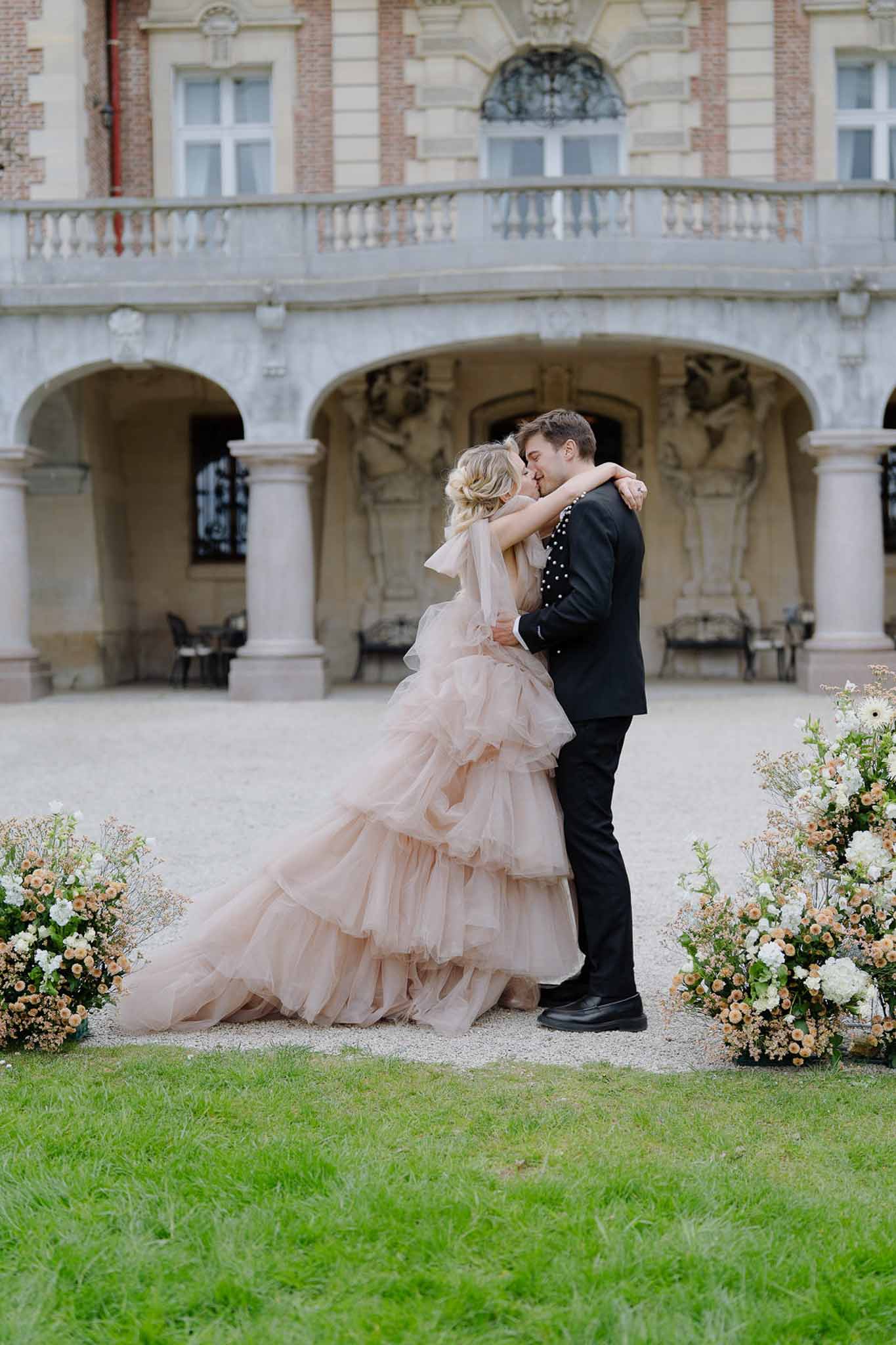 Couple kissing in chateau colonnade courtyard with peach rose and white hydrangea ground installations, bride in blush tulle