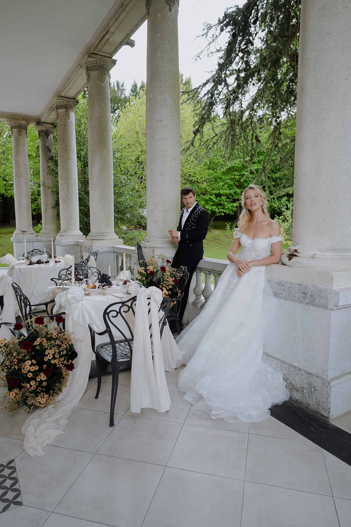 Couple on colonnaded terrace with amber chrysanthemum and burgundy rose tablescape and wrought-iron chairs