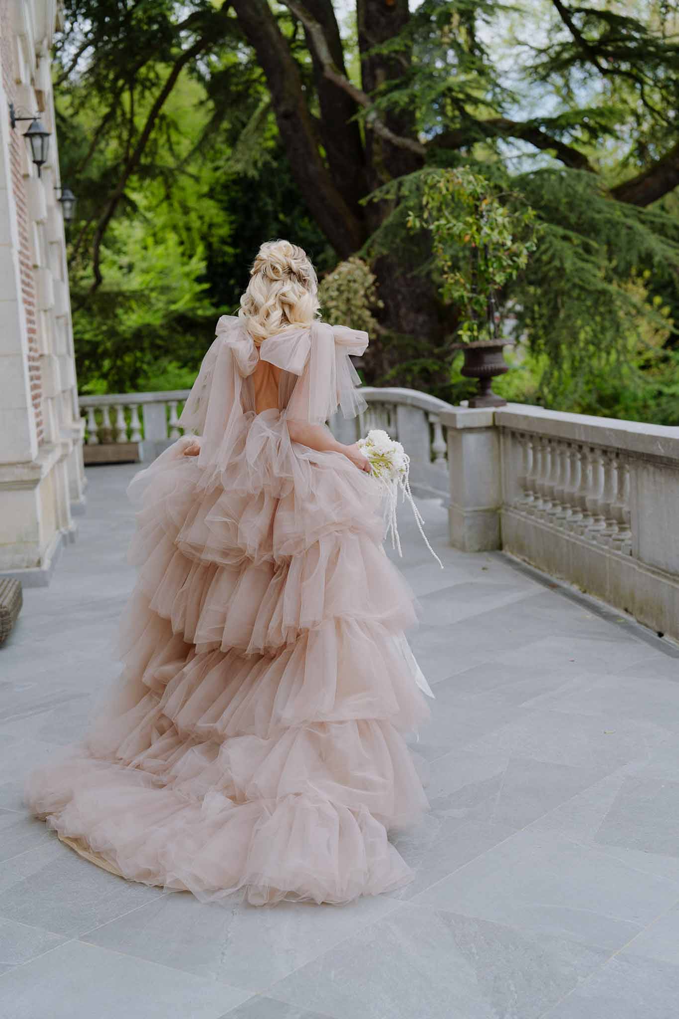 Bride from behind on chateau terrace wearing blush pink tiered tulle ball gown with dramatic train and bow detail