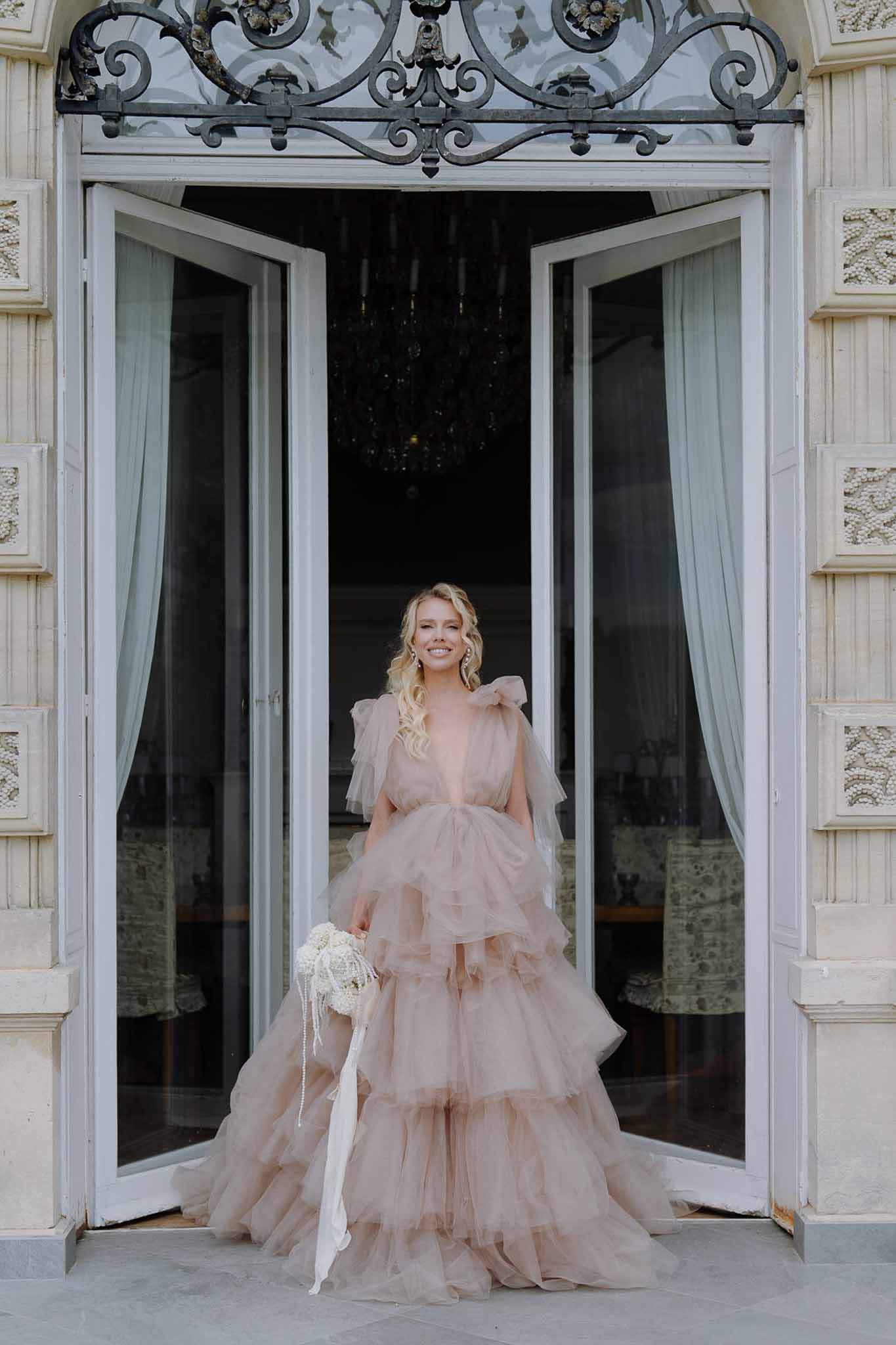 Bride in blush pink tiered tulle ball gown holding ivory bouquet at ornate chateau entrance doorway