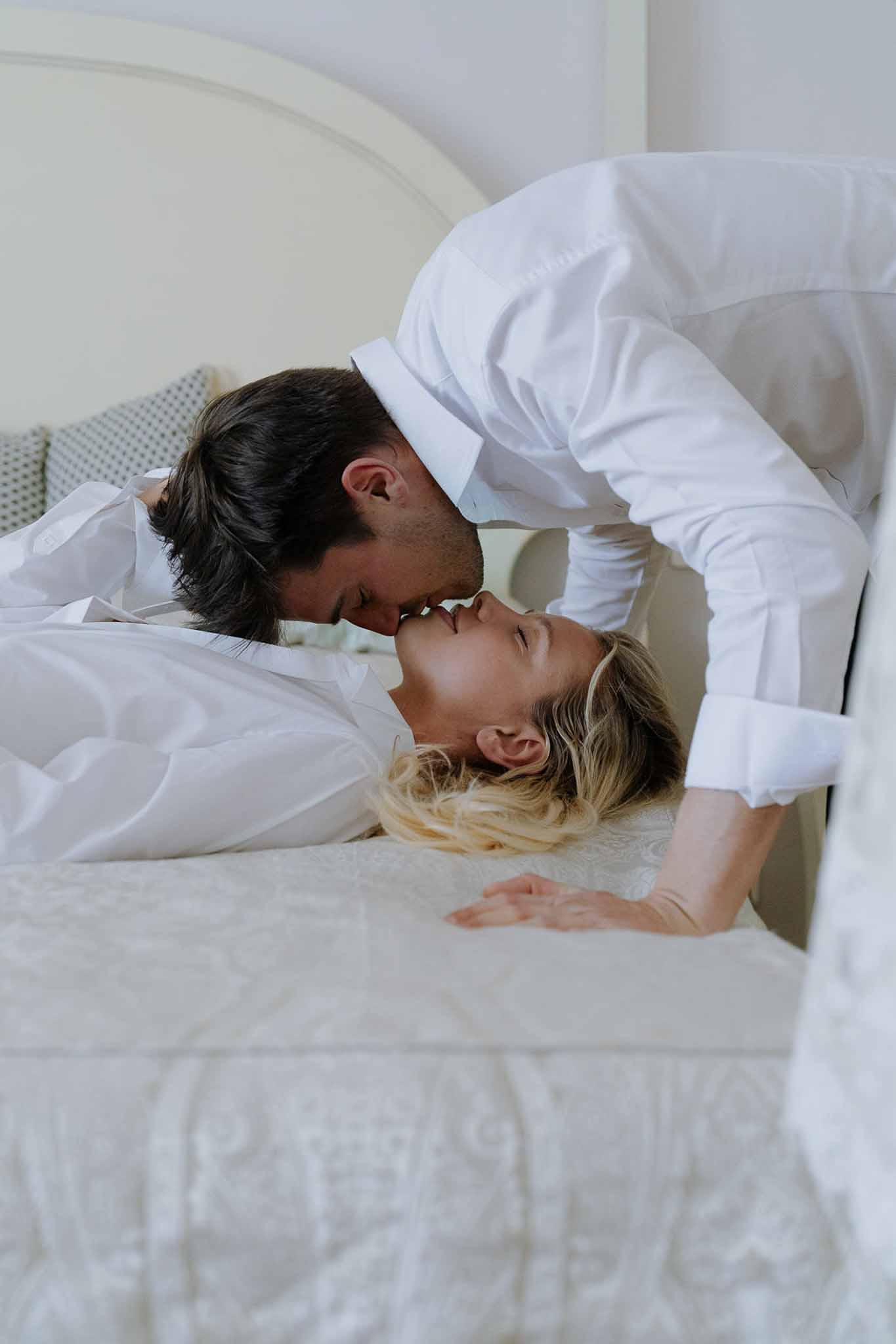 Couple kissing on white bedspread in cream-toned room during intimate getting-ready moment
