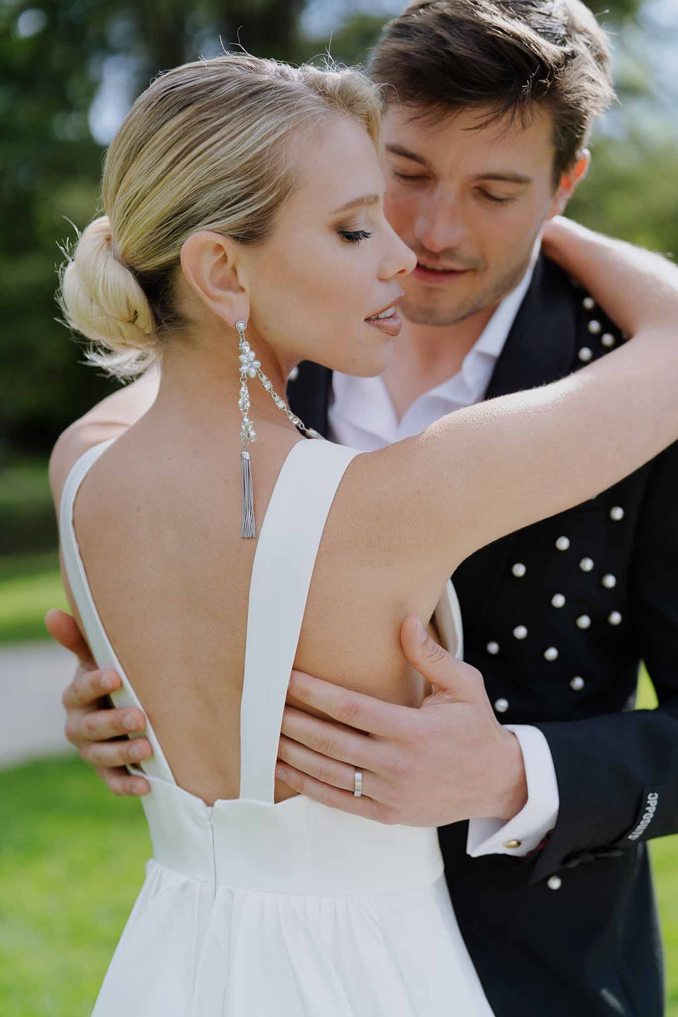 Close-up of bride's V-back gown with pearl tassel earrings and groom's embellished jacket