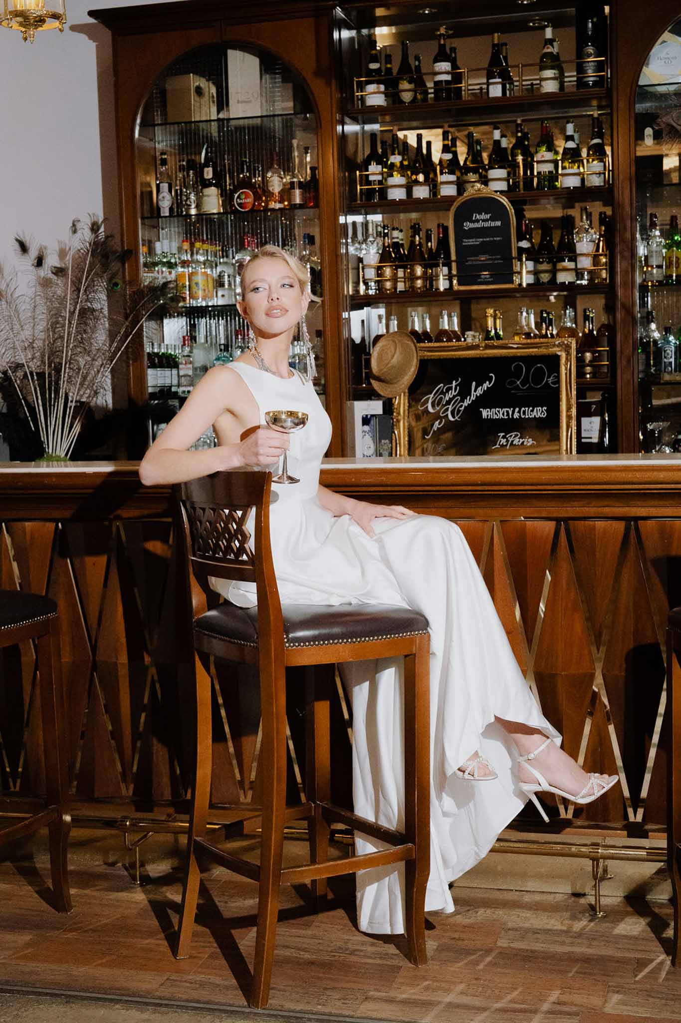 Bride in sleeveless white gown with crystal earrings seated at dark wood bar with pampas grass and spirit bottles