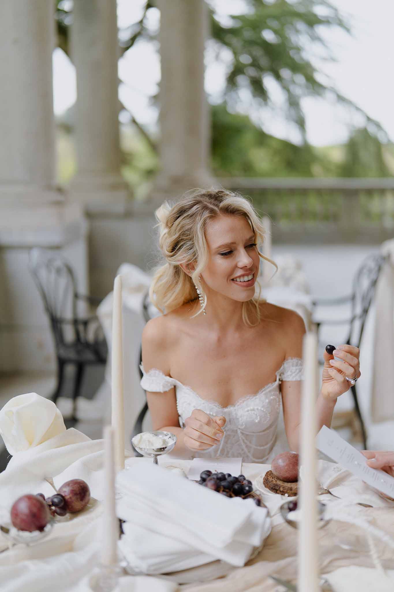 Bride in off-shoulder lace gown eating grape at styled terrace table with columns and iron chairs