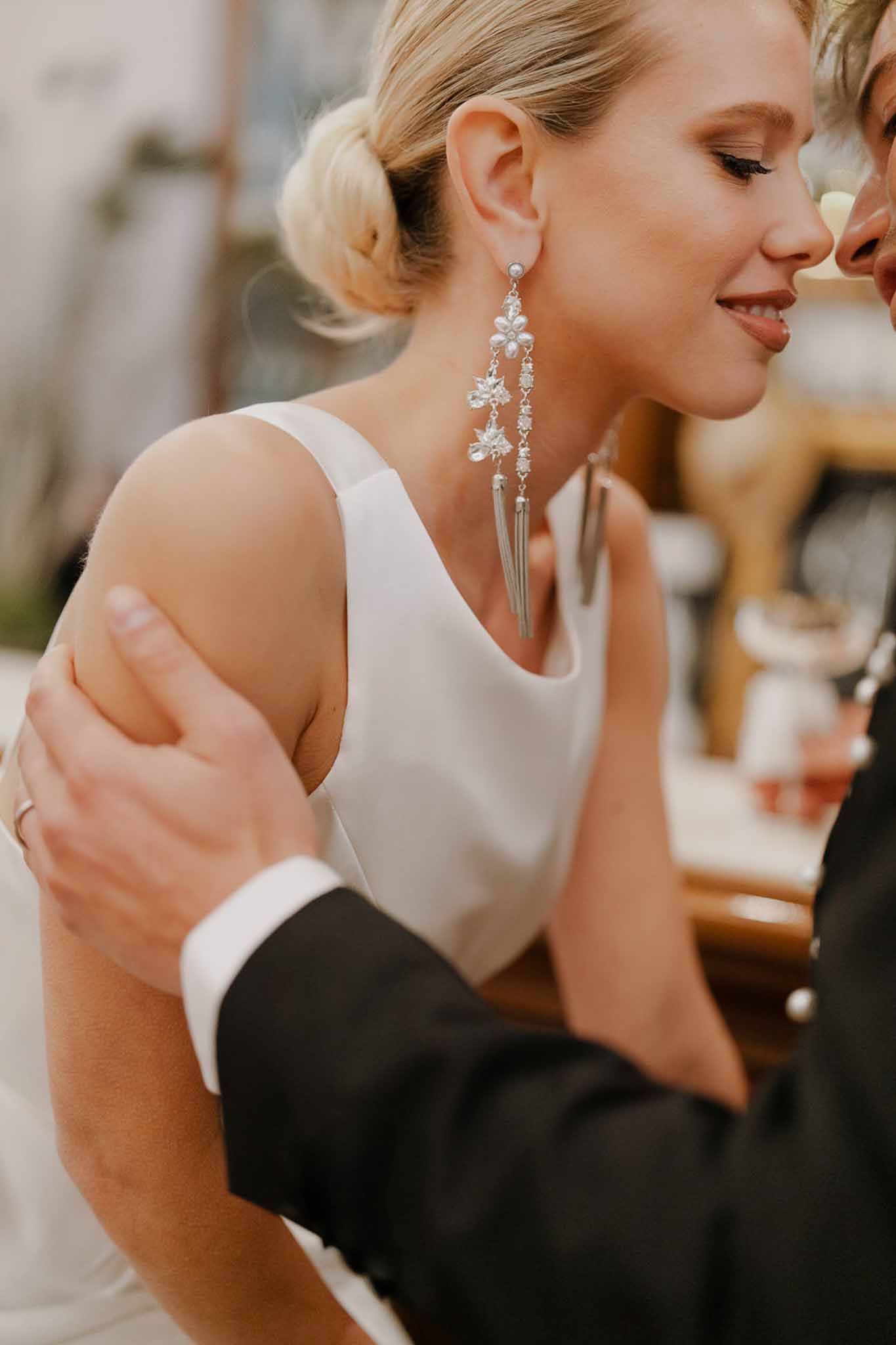 Close-up of bride with low chignon and silver chandelier earrings embraced by groom in black suit