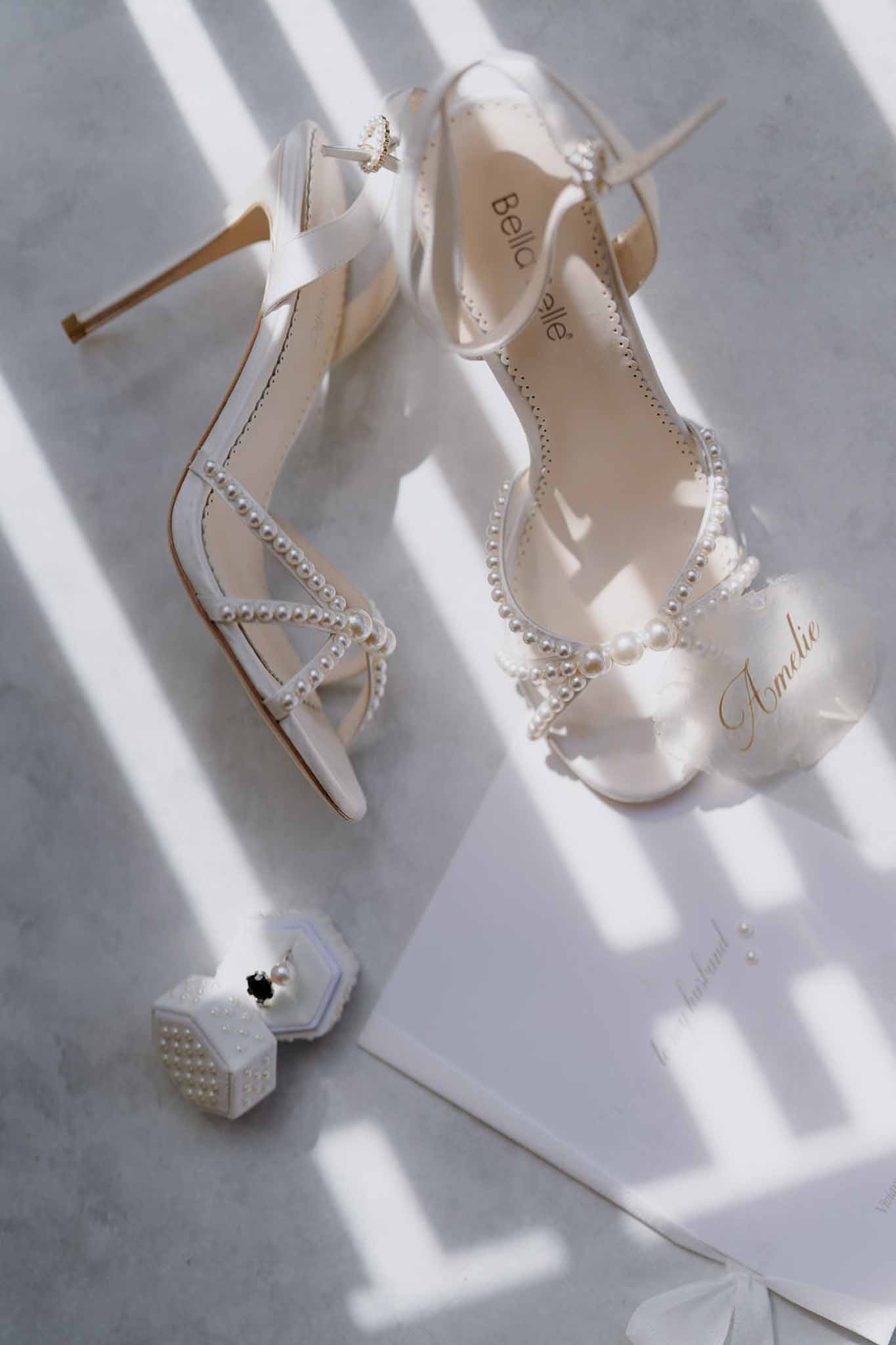 Bridal flat lay of ivory Bella Belle pearl-trimmed heeled sandals, pearl ring box, and gold script stationery on marble