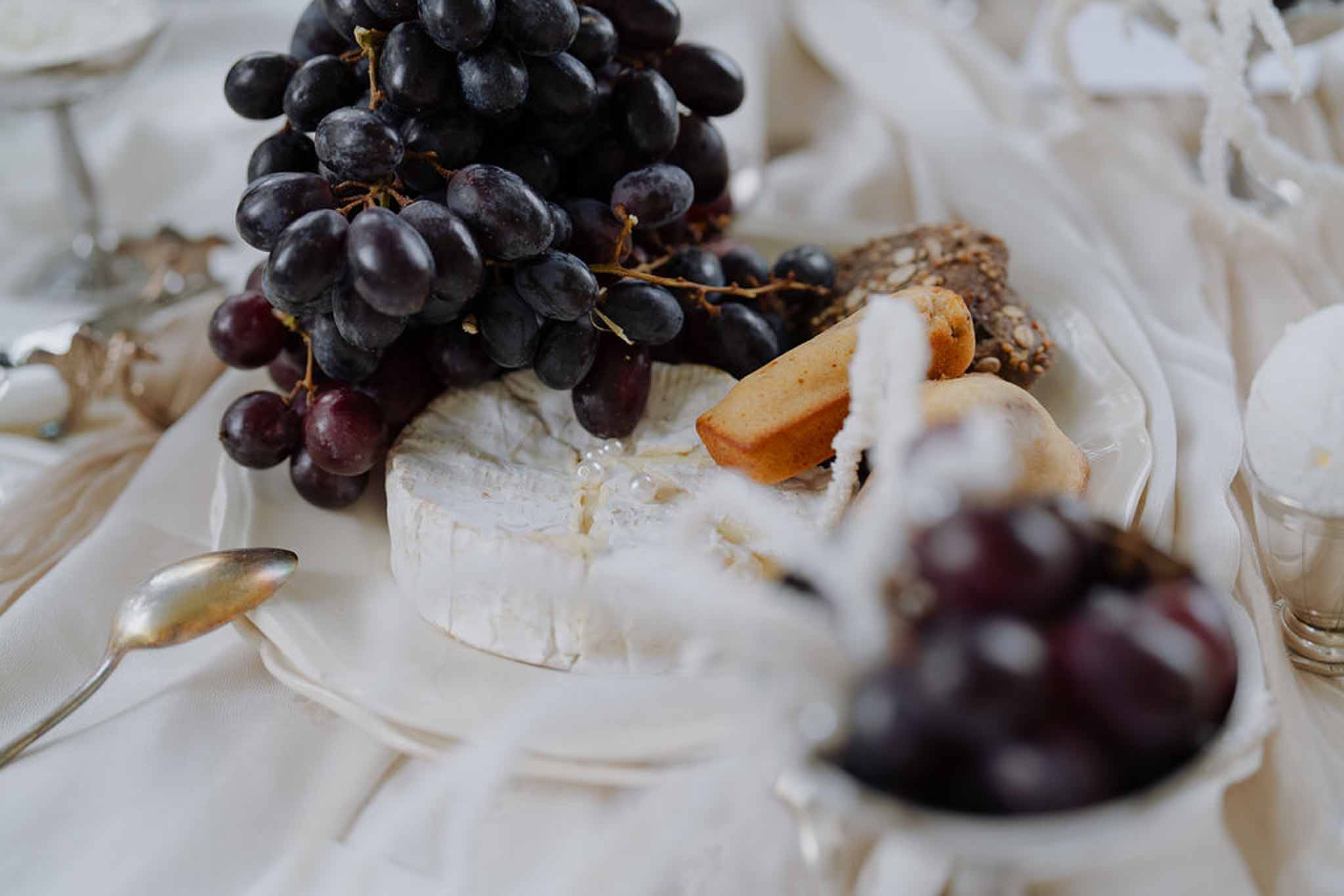 Close-up cheese board with camembert purple grapes shortbread and gold spoons on ivory linen reception table