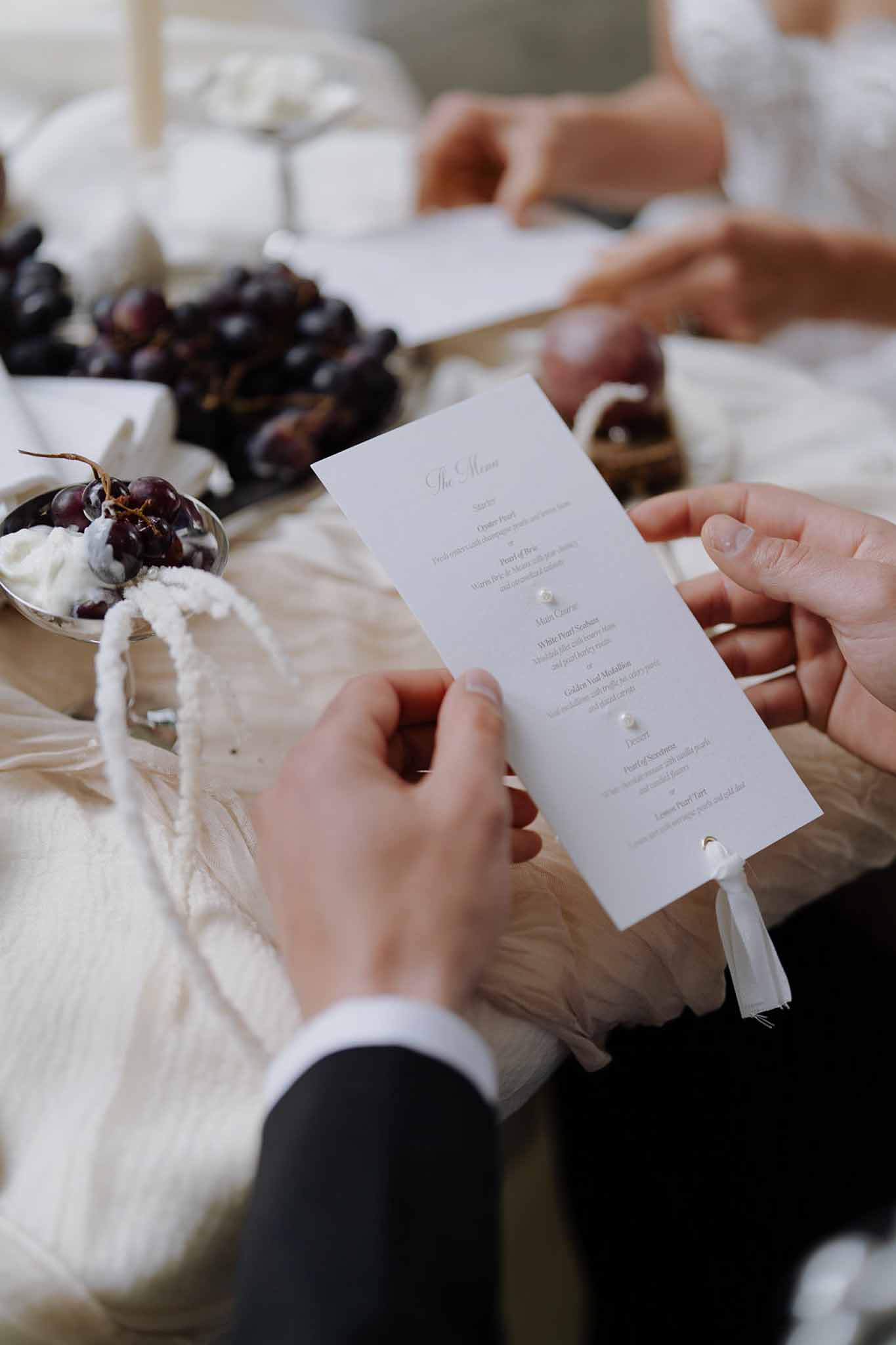Close-up of white wedding menu card with pearl embellishments held at reception table with burgundy grapes