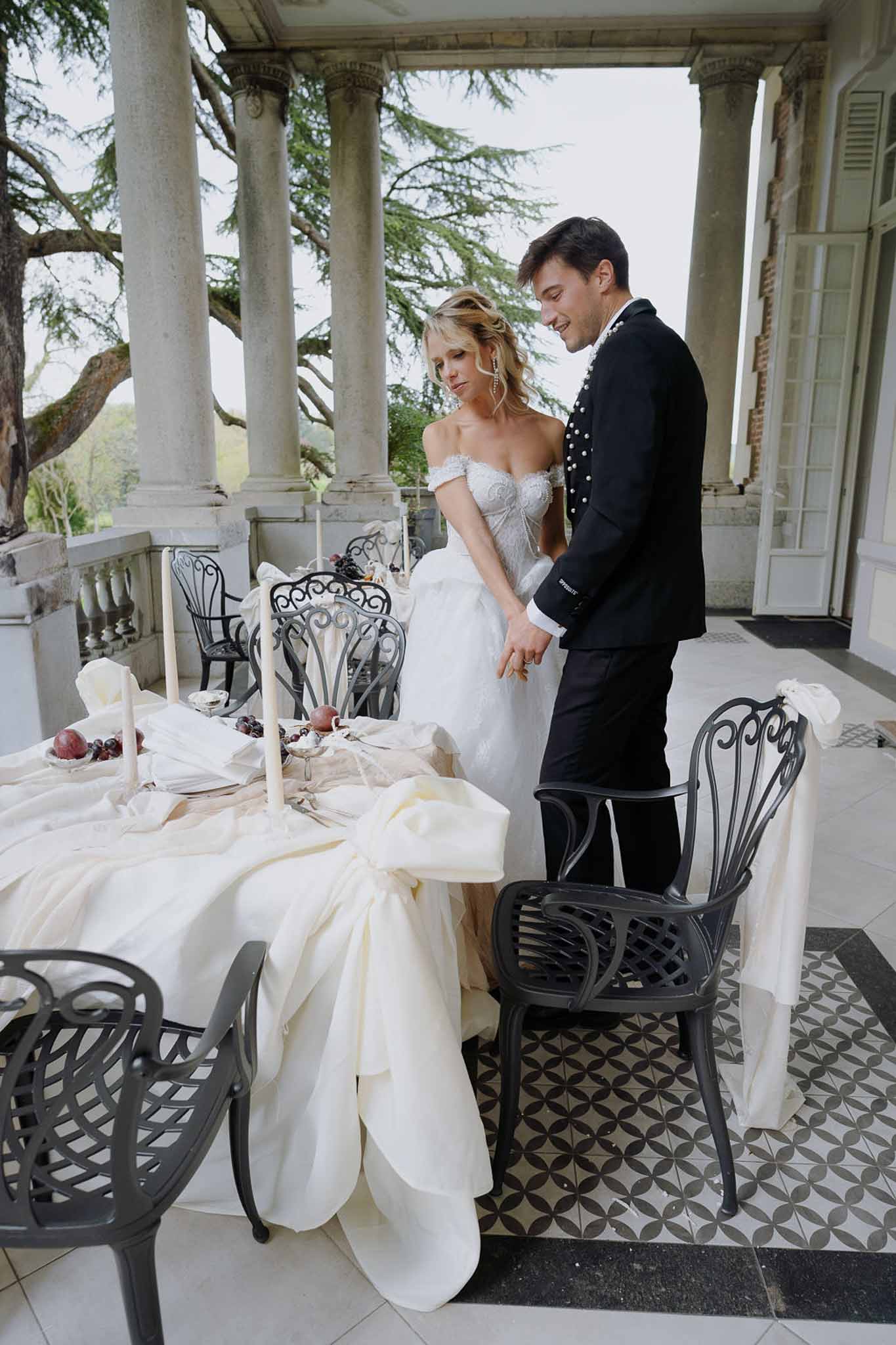 Couple on geometric-tiled chateau terrace looking at styled table with taper candles, pomegranates, and pooling linen