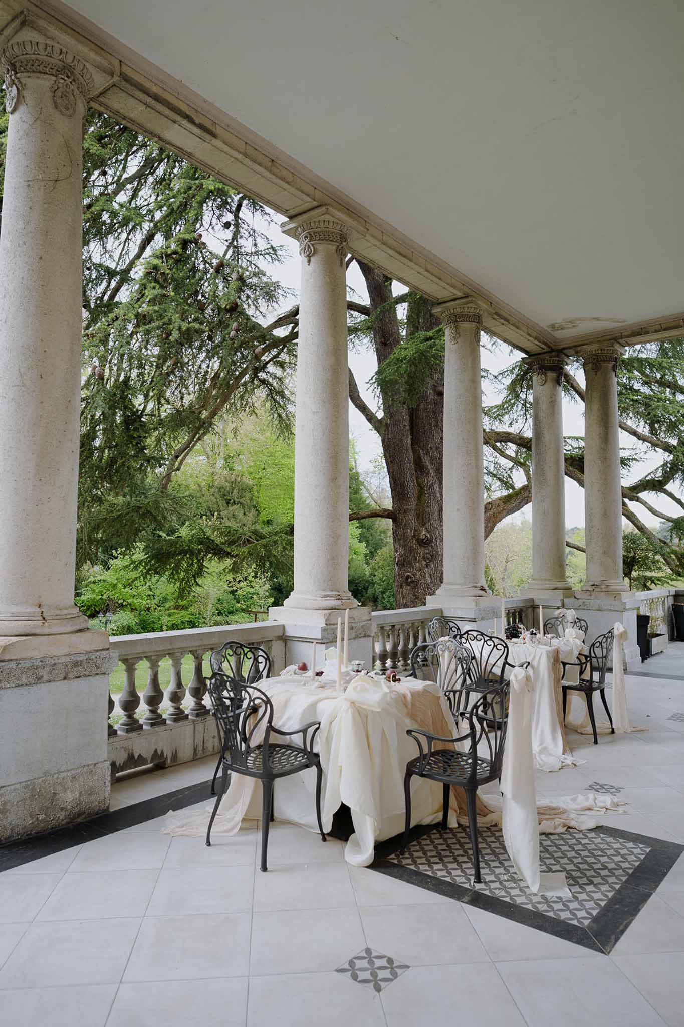 Chateau colonnade terrace with Corinthian columns, mosaic floor, iron chairs, and ivory-draped tables