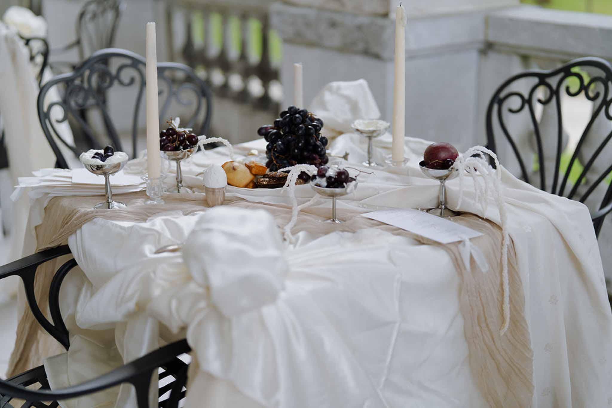 Outdoor table setting with gathered ivory linen, silver candlesticks, dark grapes, and wrought-iron chairs