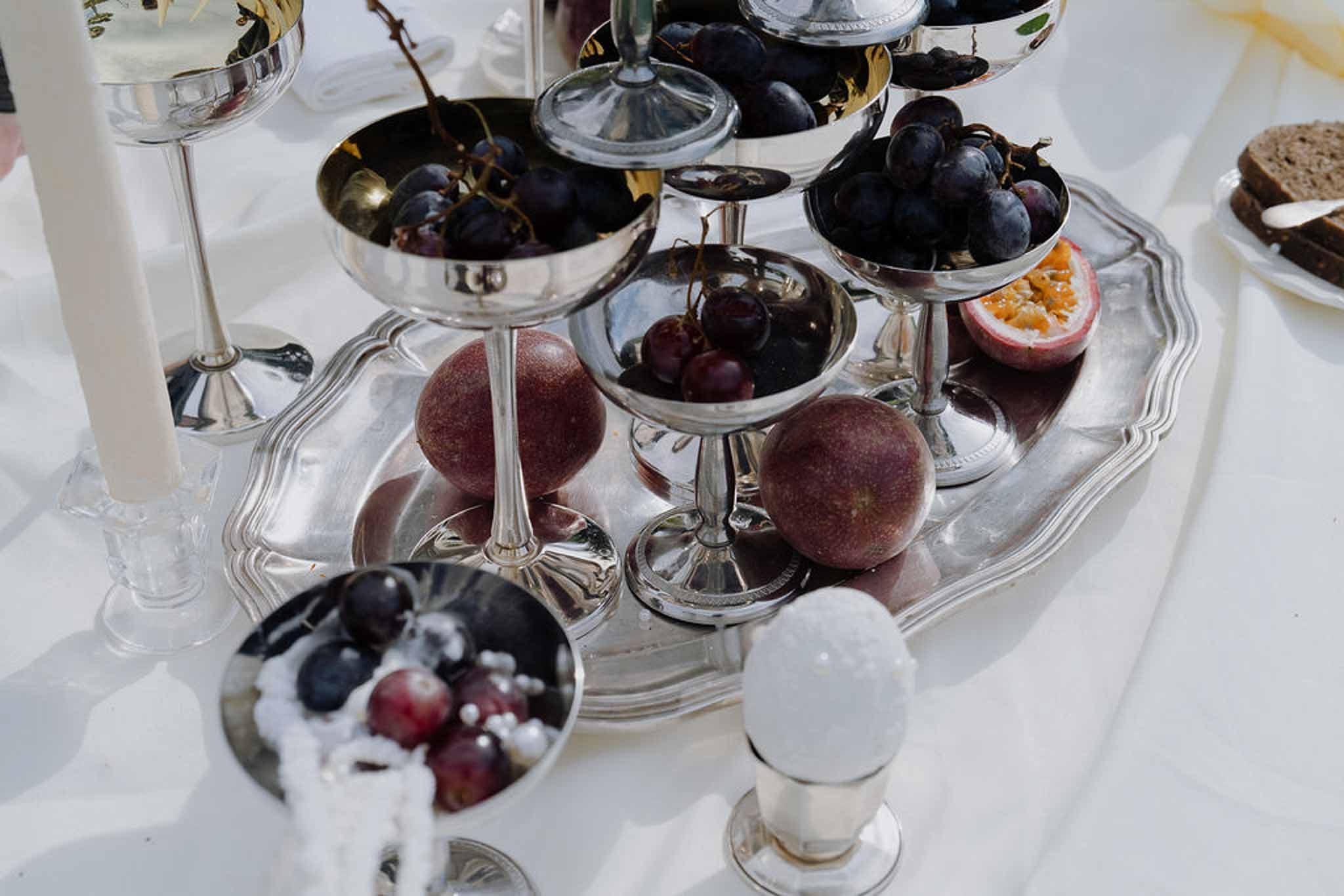 Silver tiered tray with dark grapes, cherries, and passion fruits beside taper candle on white linen
