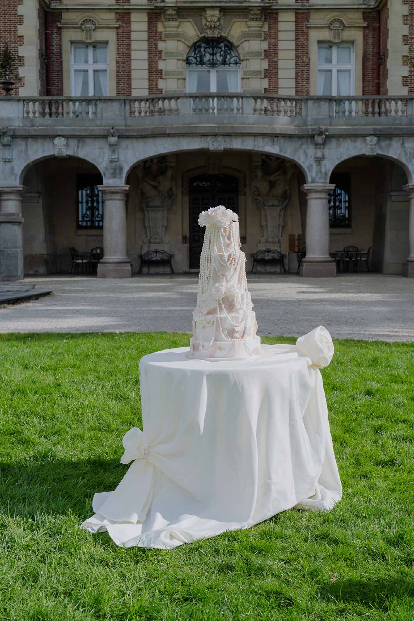 Multi-tiered blush cake with pearl strands and sugar bows on lawn before chateau with stone arched colonnades