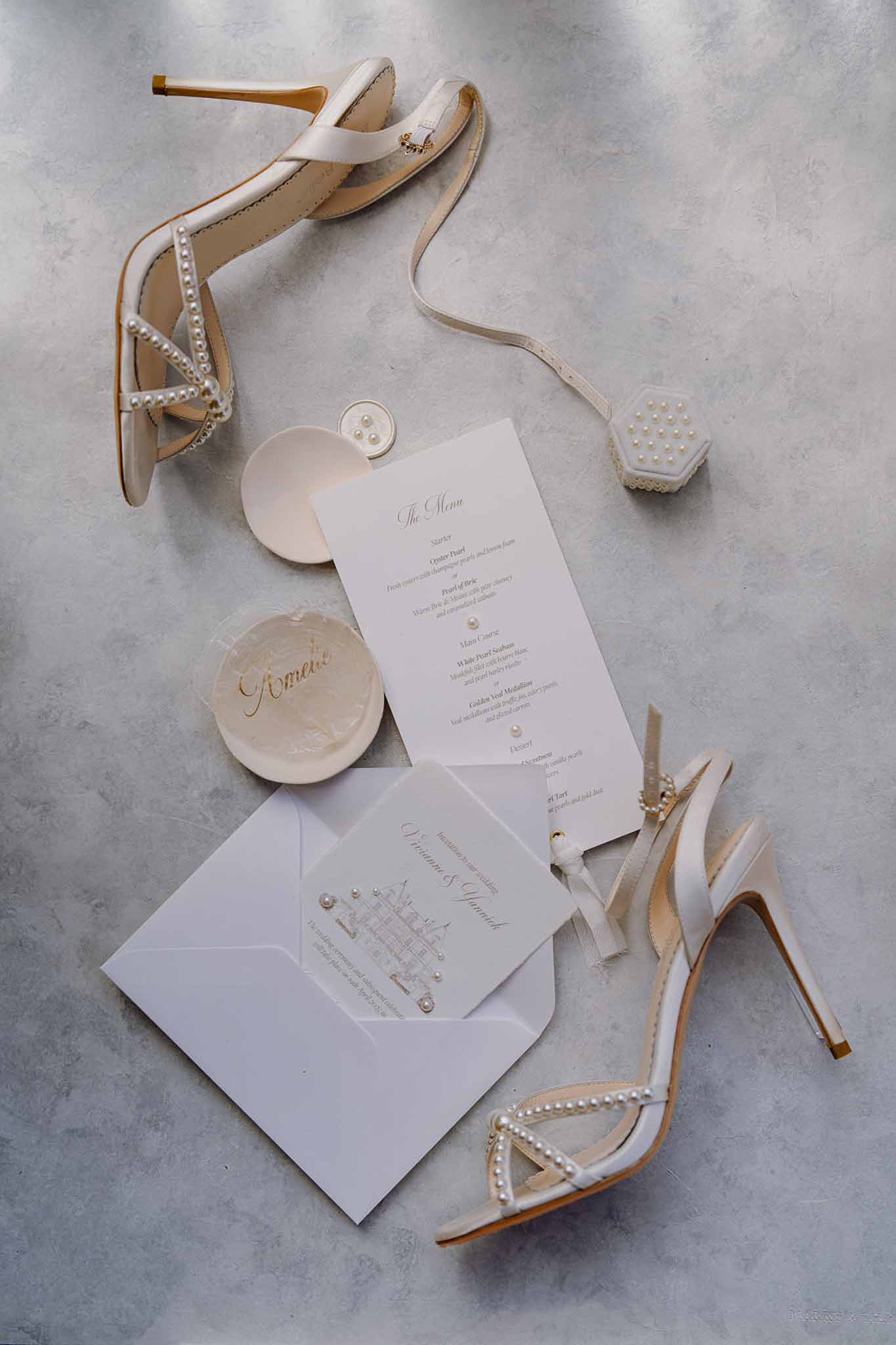 Bridal flat lay with pearl-strap heels, calligraphy invitation suite, and rings on grey marble surface