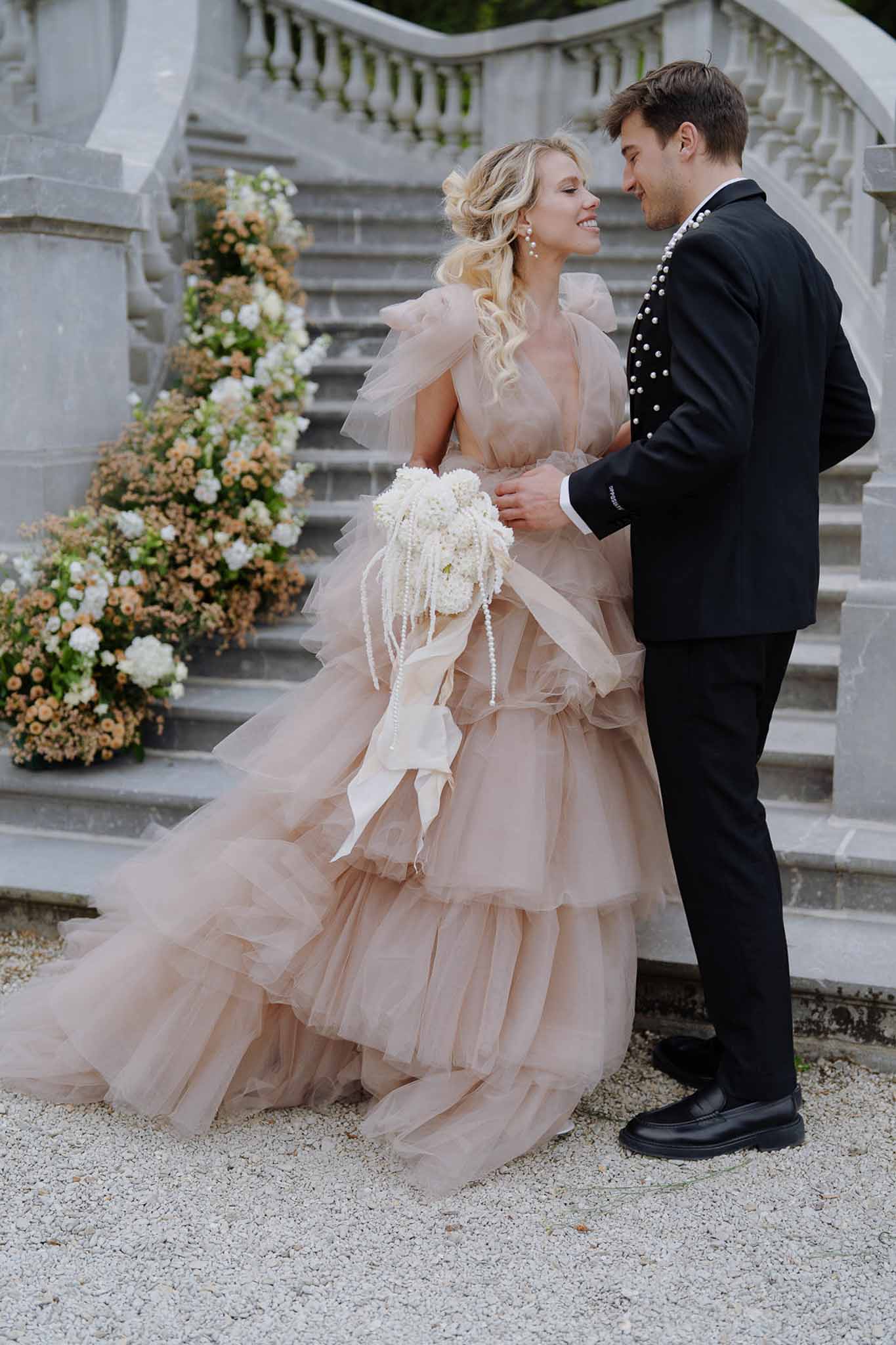 Couple at stone staircase bride in blush tiered tulle gown with pearl bouquet groom in pearl-embellished tuxedo