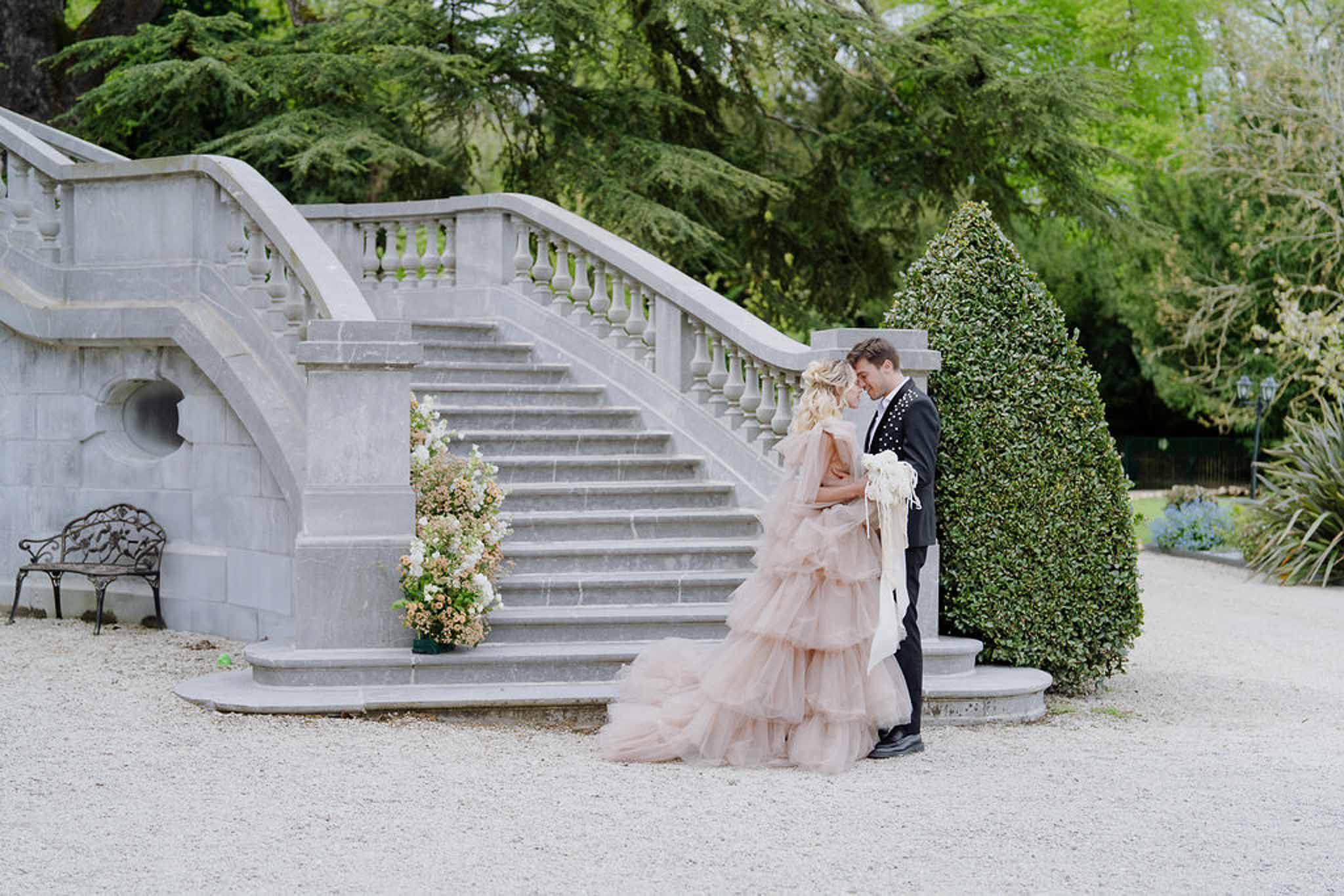 Bride in blush tulle gown and groom in tuxedo standing before grand stone double staircase at chateau