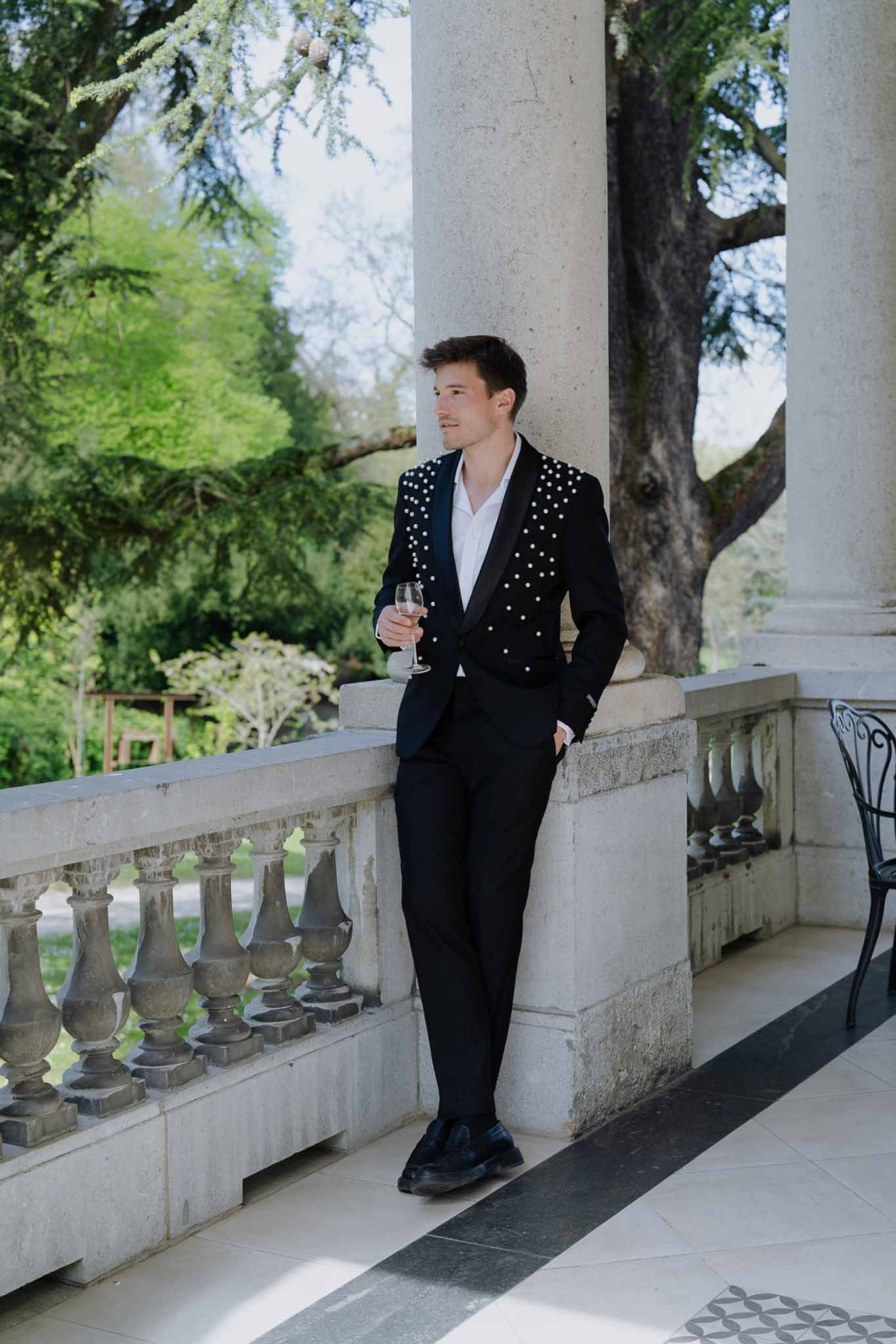 Groom leaning on stone column holding champagne in pearl-studded tuxedo jacket on chateau balustrade terrace