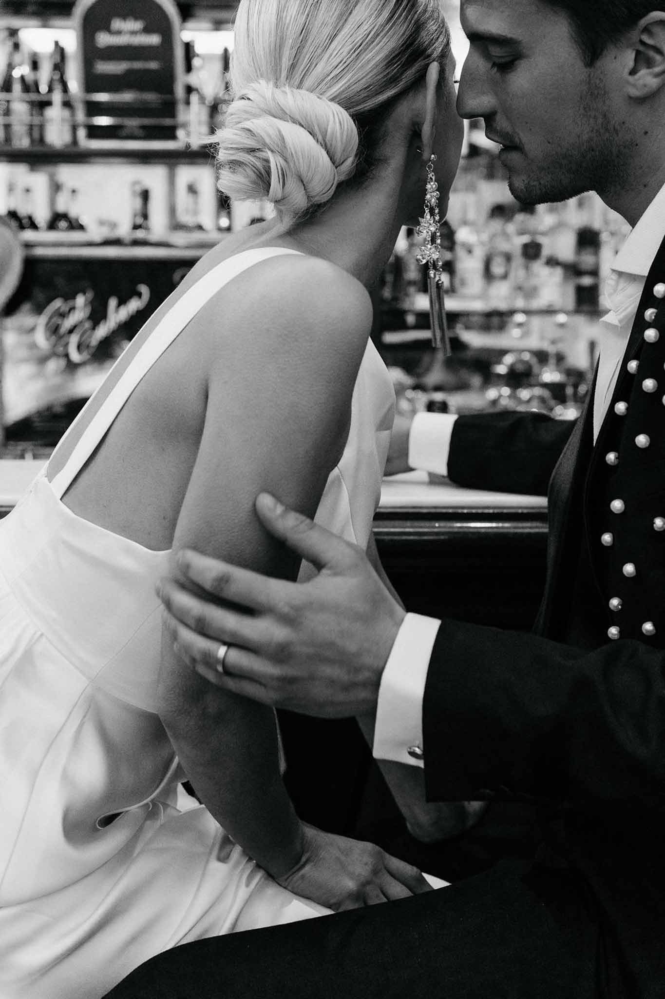 Black and white close-up of bride and groom in intimate moment at indoor bar, bride in low-back halter dress