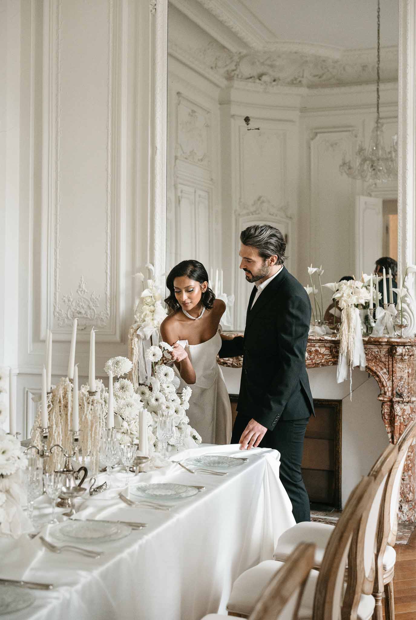 Bride and groom reviewing reception table settings in neoclassical hall with white floral arrangements