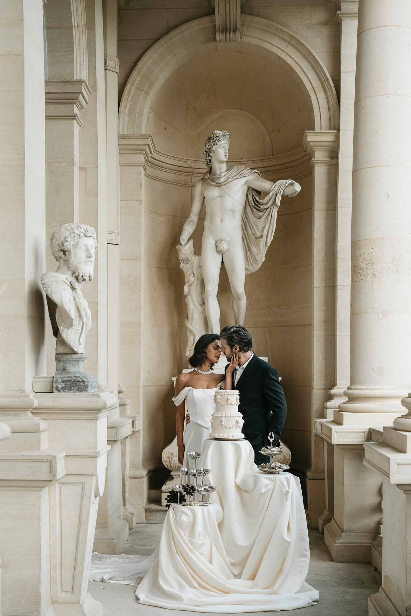 Couple cutting wedding cake in neoclassical gallery with marble columns and classical statuary