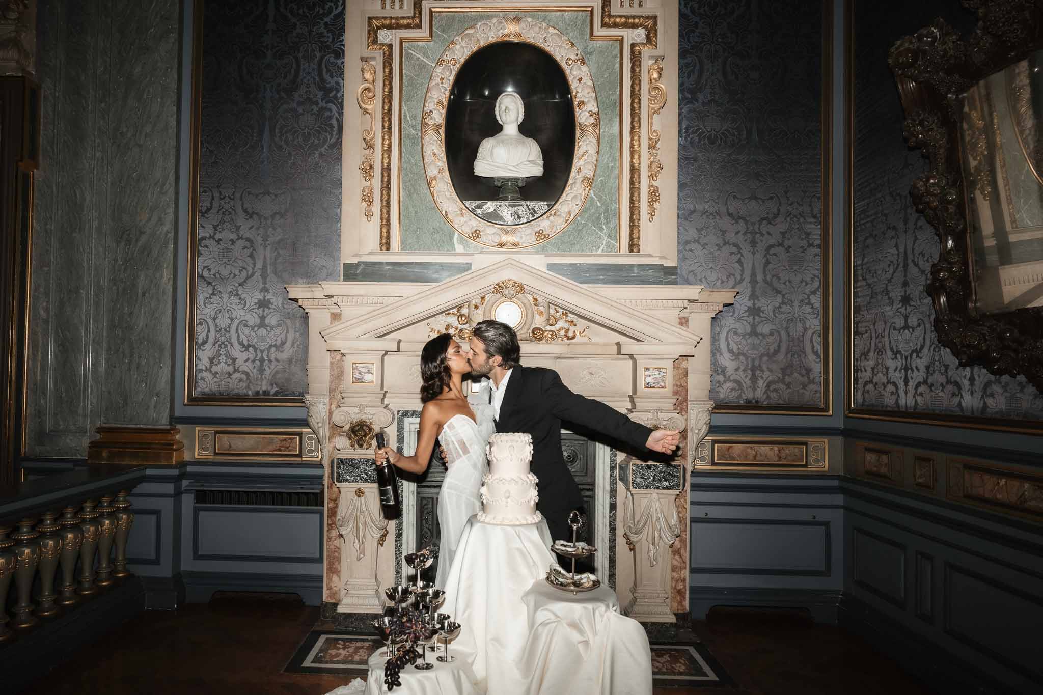 Bride and groom kissing during reception in grand neoclassical interior with wedding cake and candelabras