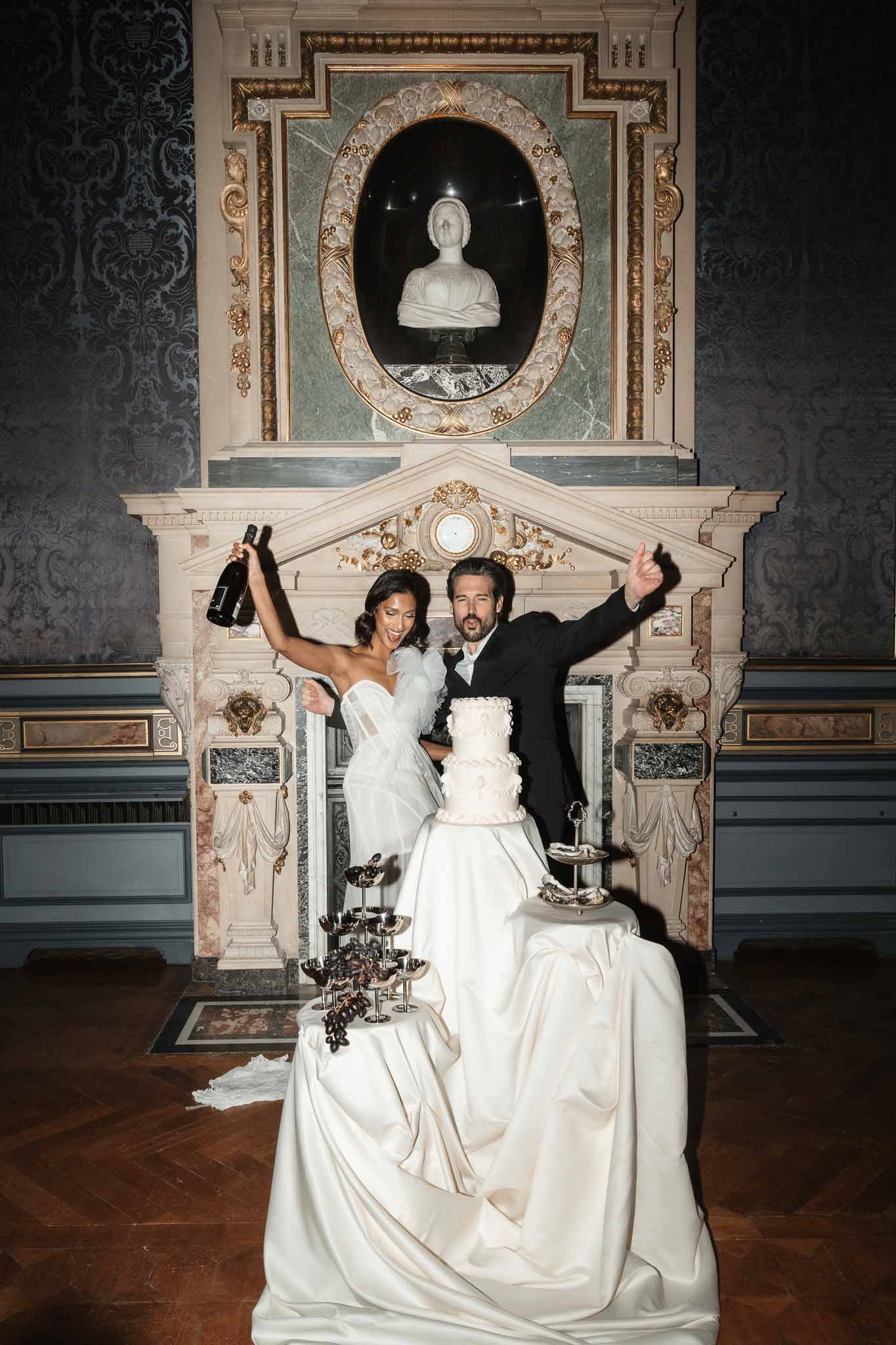 Bride and groom celebrating with champagne and wedding cake at elegant marble fireplace reception venue