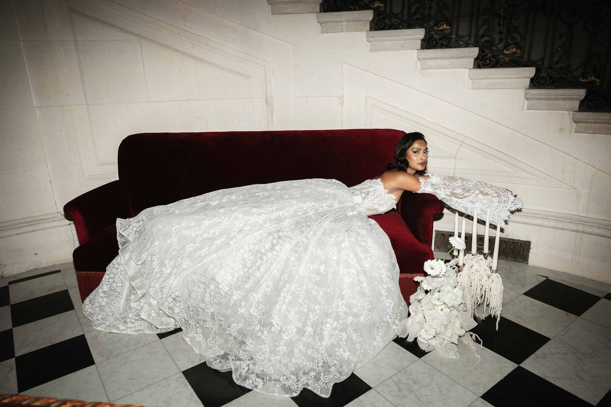 Bride in ivory lace gown posing on burgundy velvet sofa in elegant interior with checkered marble floors