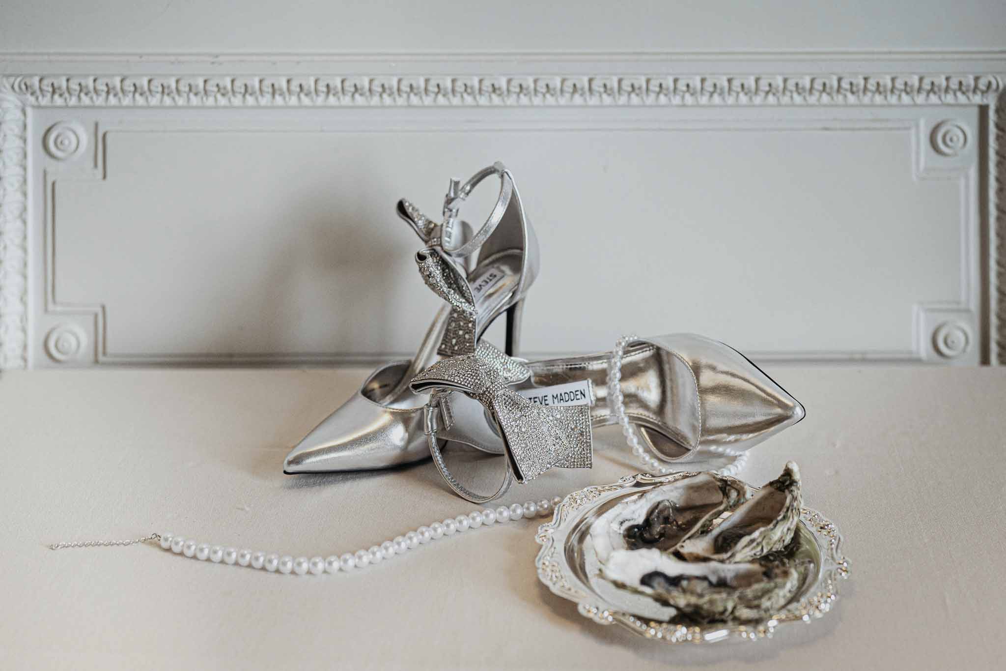 Bridal accessories flat lay with silver heels, pearl necklace and earrings on white molding background
