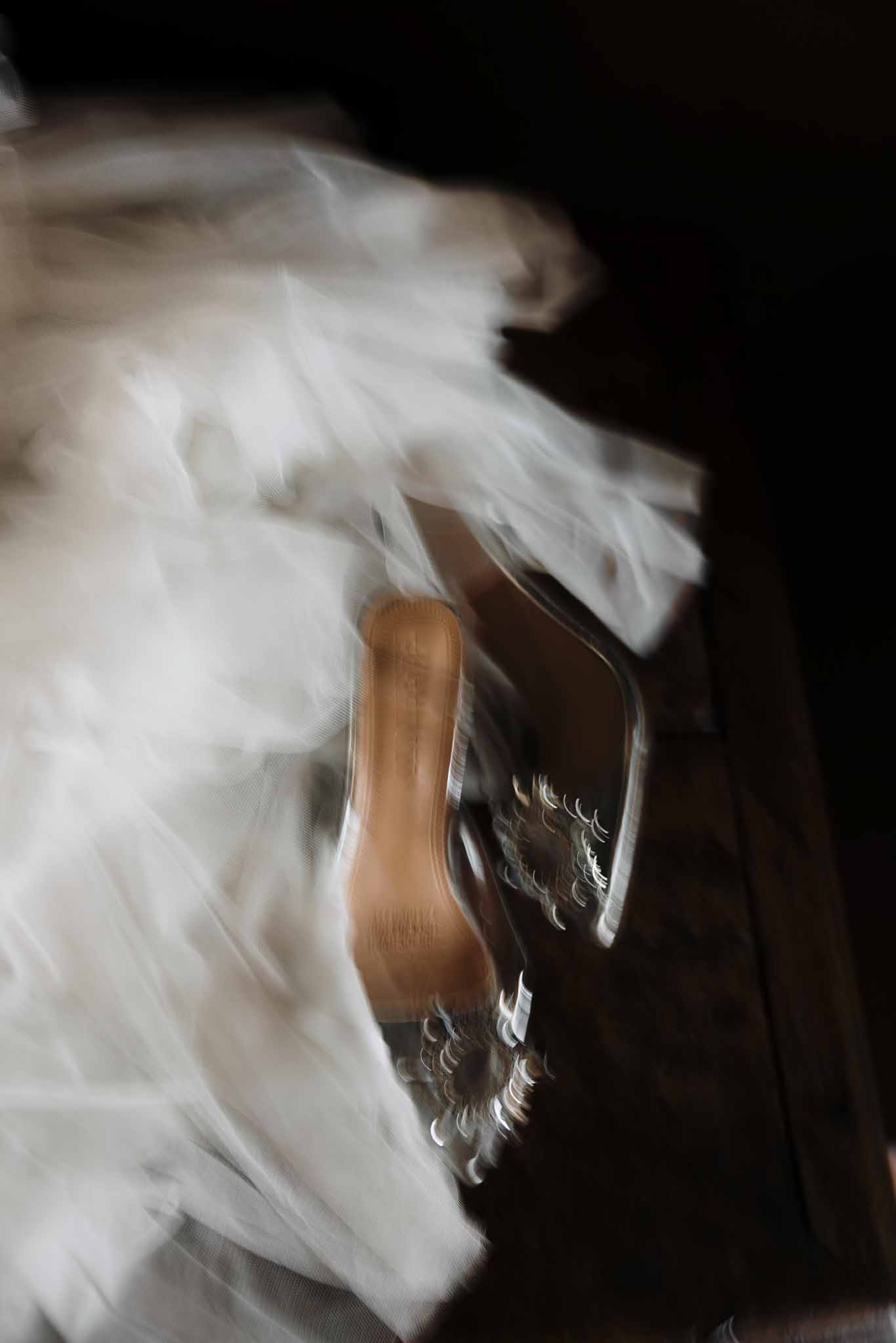 Motion-blurred clear crystal mules with white tulle veil layers on dark wooden surface in artistic detail shot