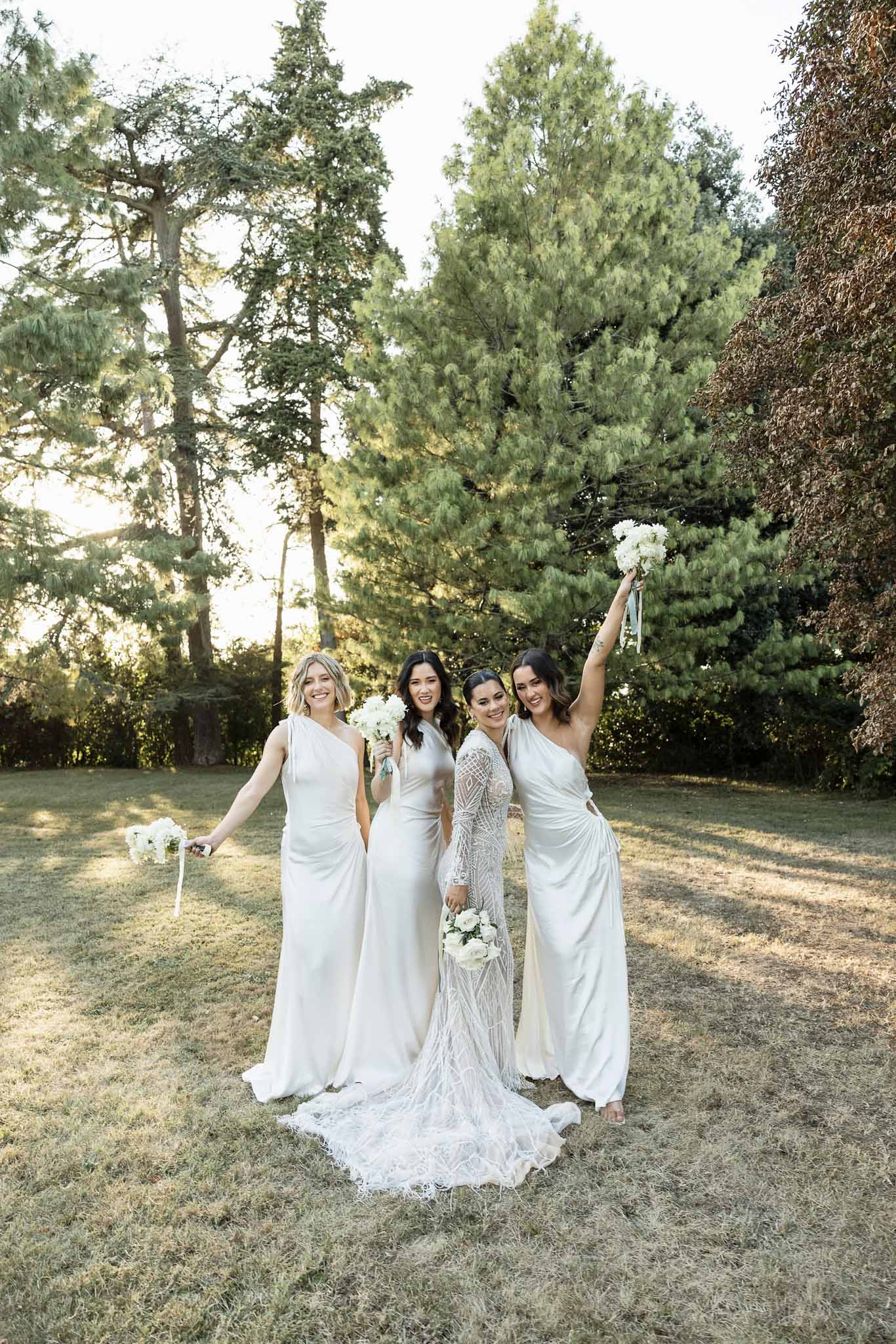 Bride and three bridesmaids in ivory satin dresses laughing together on a sunlit lawn holding white rose bouquets